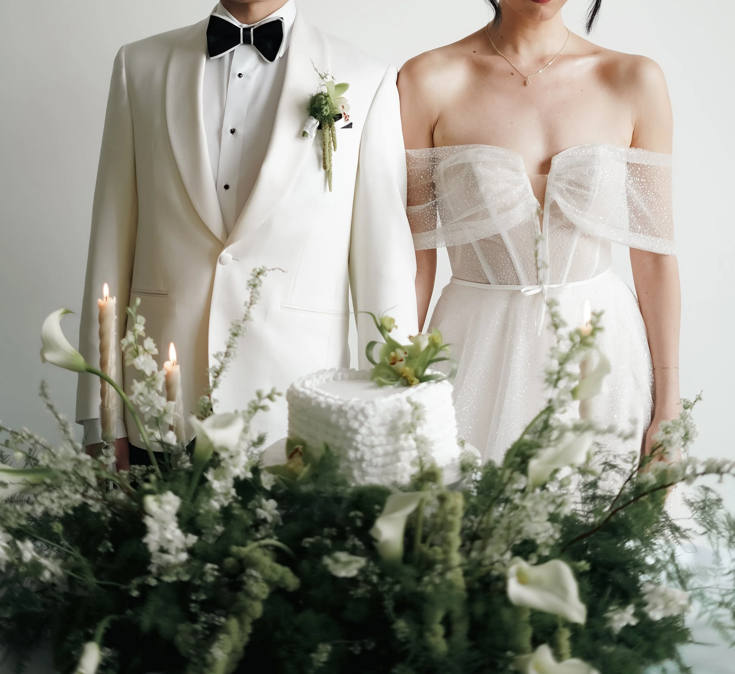 A bride and groom at their wedding, dressed in white, with the groom wearing a tuxedo with a black bow tie and boutonniere, and the bride wearing a strapless, off-shoulder wedding gown with sheer polka dot detailing.