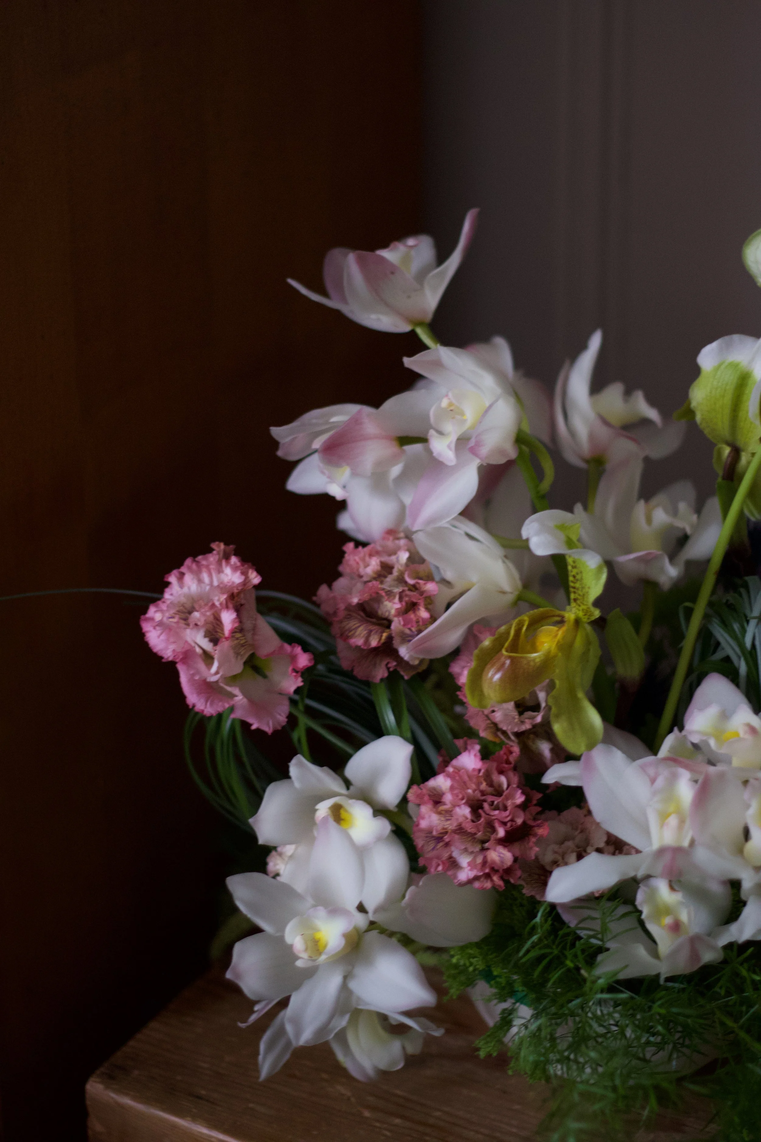 A floral arrangement with white, pink, and green flowers on a wooden table, set against a dark background.