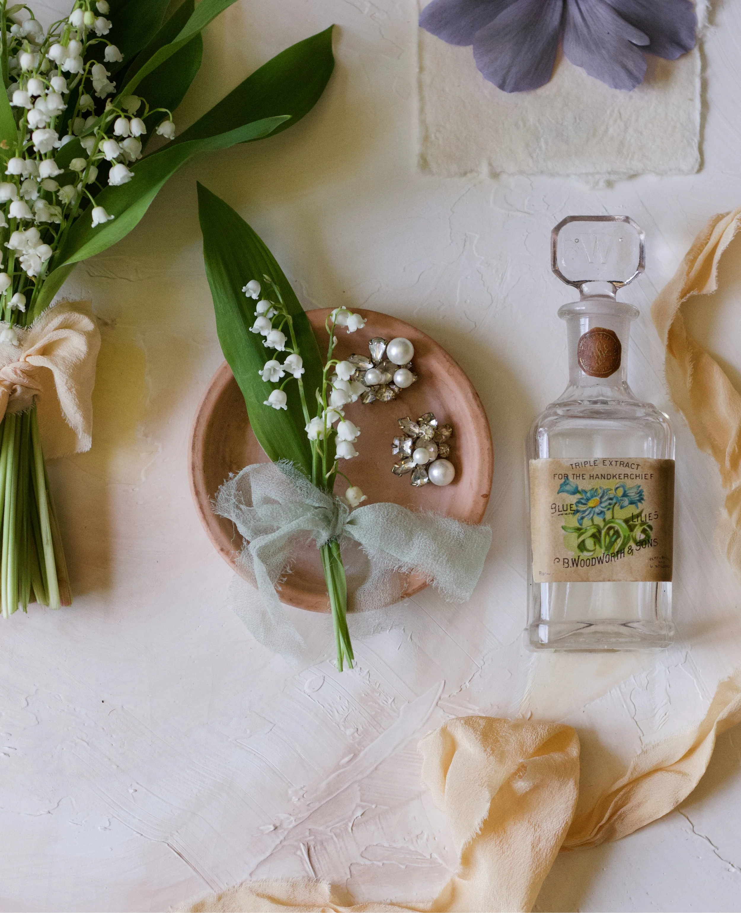 A small bouquet of lily of the valley flowers with green leaves, tied with a light green ribbon, placed on a pink dish. Next to it are vintage-style pearl and rhinestone earrings. To the right, there is a clear glass perfume bottle with a label and a