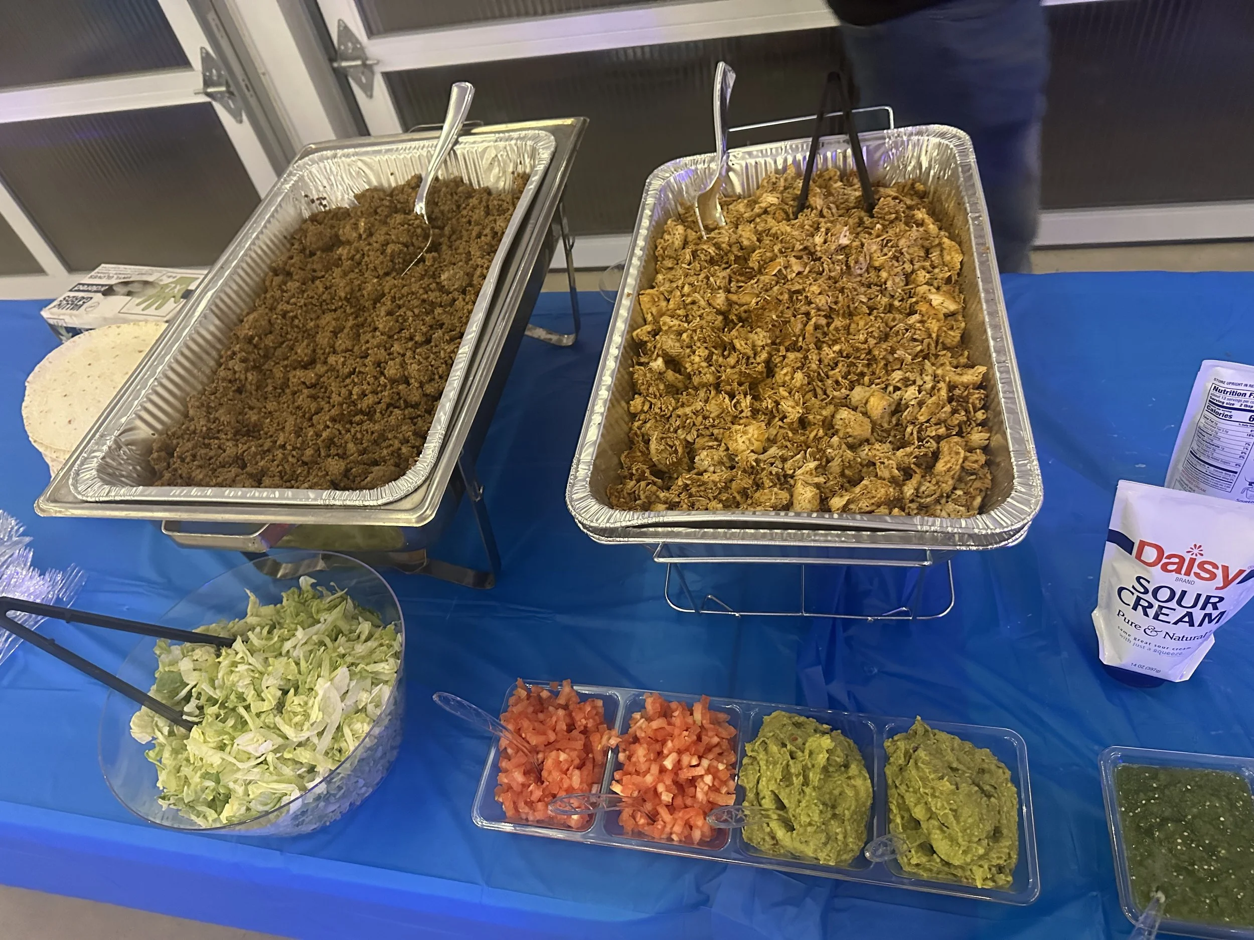 Buffet table with trays of seasoned ground beef and shredded chicken, bowls of chopped lettuce, diced tomatoes, guacamole, and salsa, with sour cream and tortilla chips on the side.