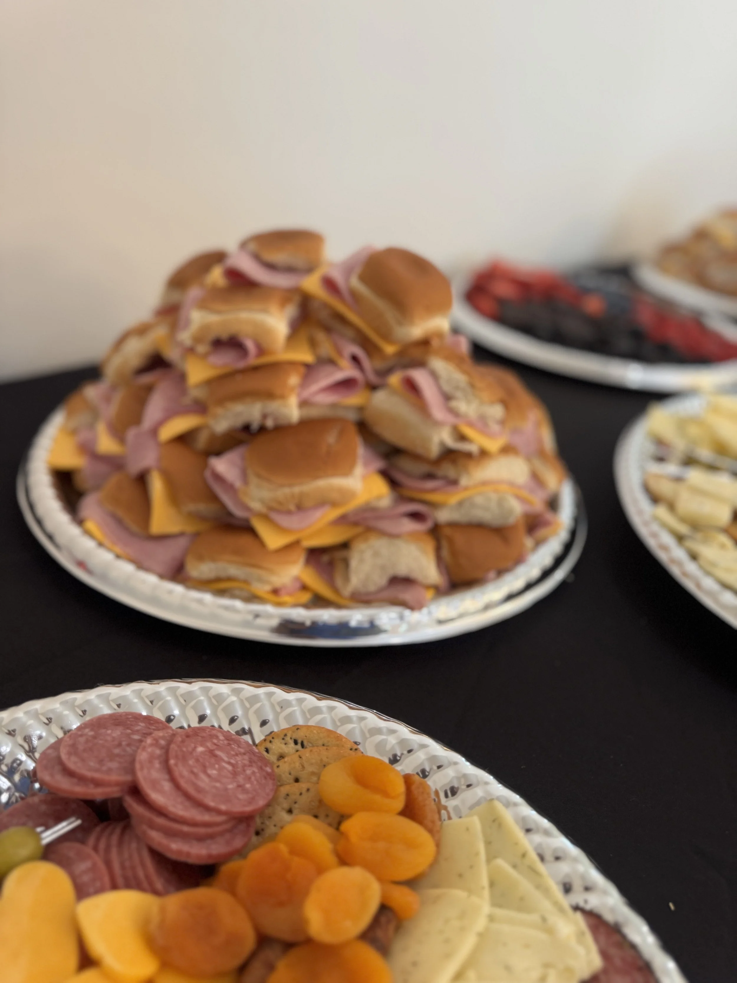 A platter of mini sandwiches stacked in a pyramid with ham and cheddar cheese on small buns. In the foreground, a platter of assorted cold cuts, sliced cheese, and dried apricots. Other platters of food are visible in the background.
