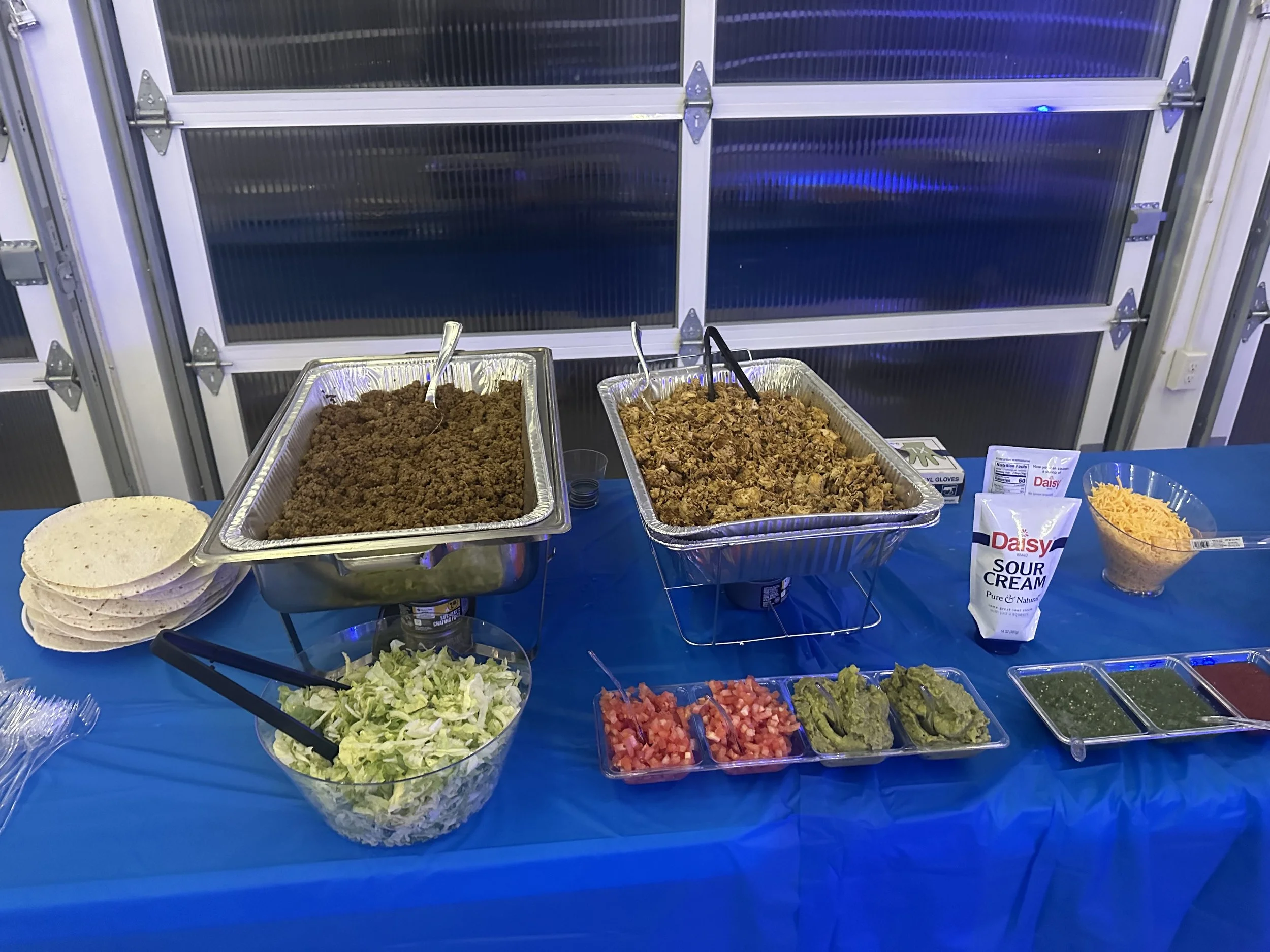 A buffet table with trays of seasoned ground beef and shredded chicken, bowls of shredded cheese, sour cream, chopped tomatoes, sliced avocados, and salsa, along with tortillas and utensils.