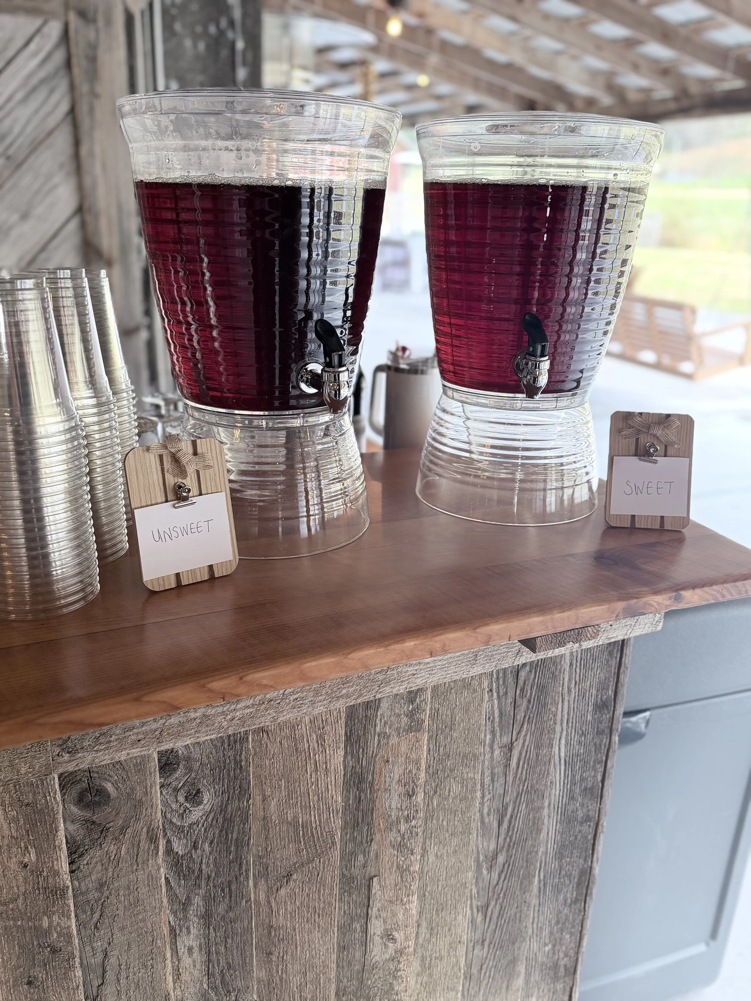 Two large glass beverage dispensers filled with dark purple juice, with spigots at the bottom, placed on a wooden counter. To the left, there are stacked clear plastic cups. Small wooden signs with labels read "Unsweet" and "Sweet" in front of each d