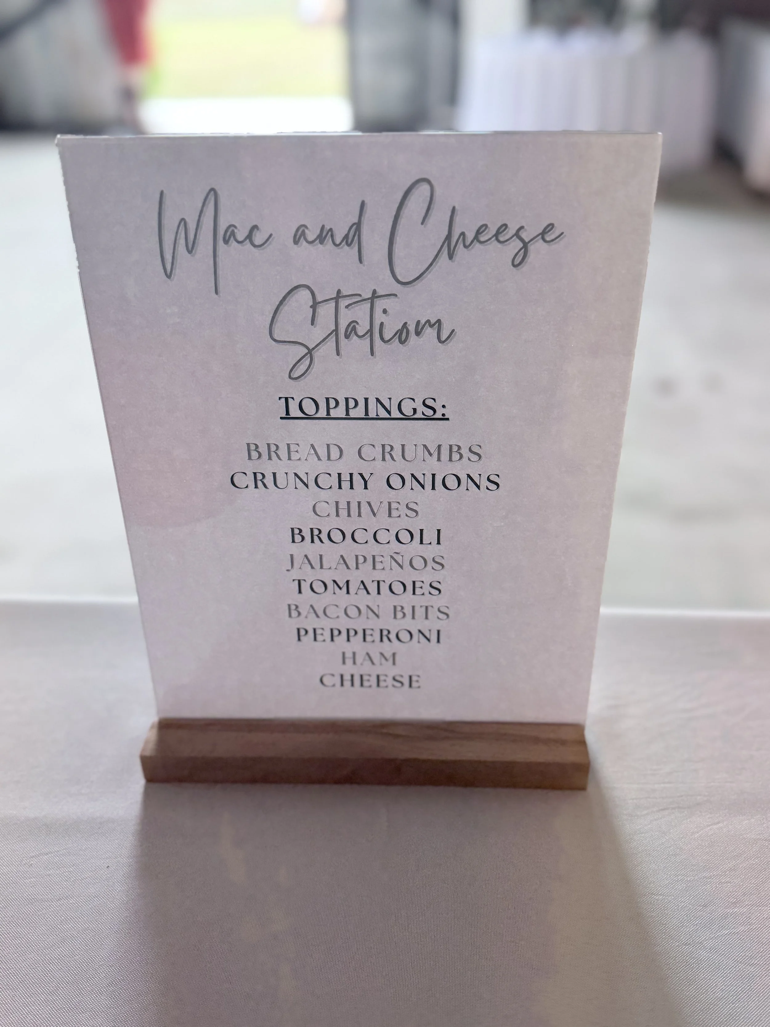 Menu sign for Mac and Cheese Station listing toppings such as bread crumbs, crunchy onions, chives, broccoli, jalapenos, tomatoes, bacon bits, pepperoni, ham, and cheese.