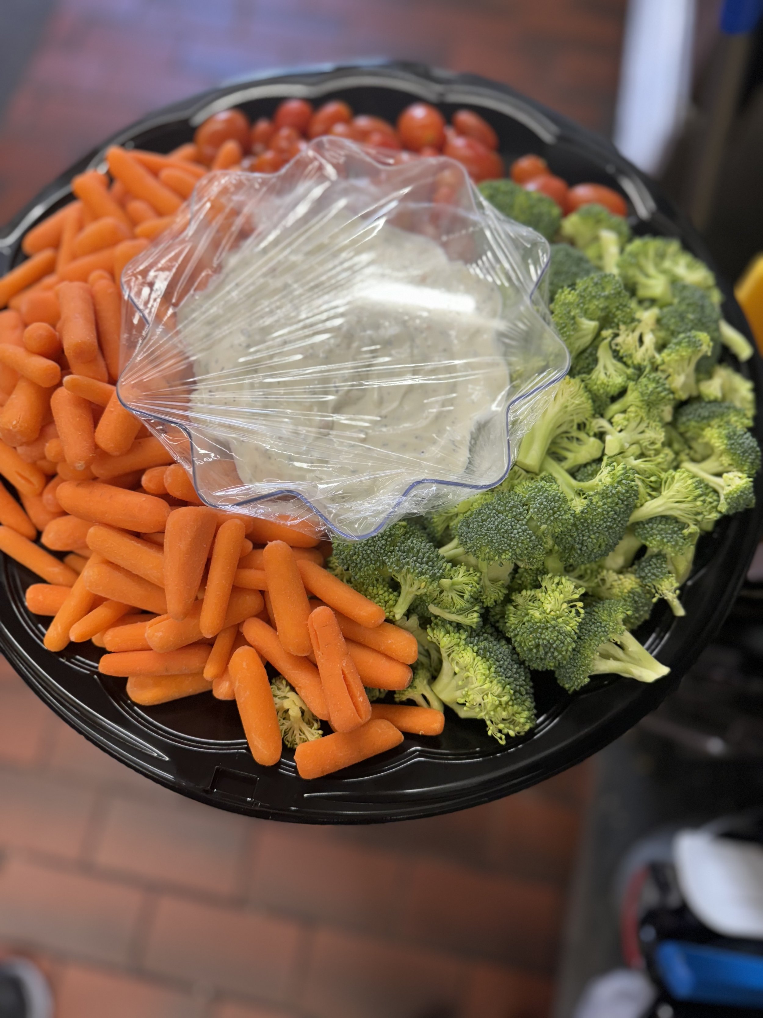 A black platter filled with broccoli florets, baby carrots, and grape tomatoes, with a small container of ranch dressing on top.