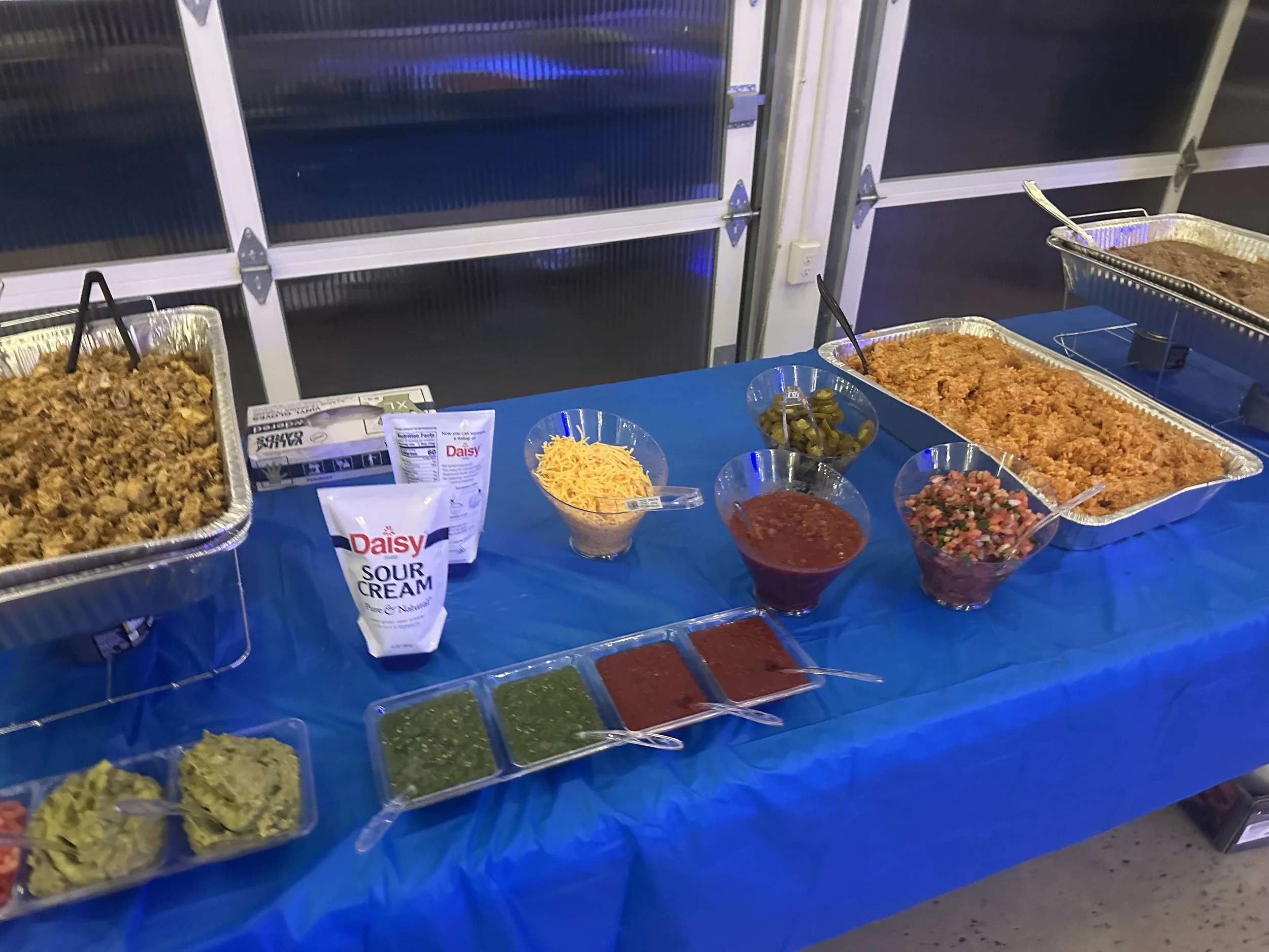 A buffet table with trays of cooked Mexican rice and shredded meat, bowls of shredded cheese, jalapeños, salsa, and chopped tomatoes, along with bowls of sour cream, guacamole, and various salsas. The table is covered with a blue tablecloth.