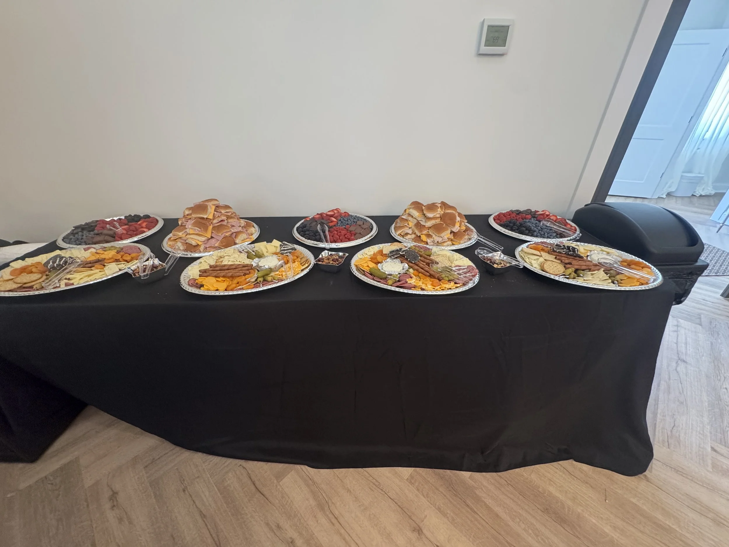 A long rectangular table with a black tablecloth holds trays of breakfast foods and snacks, including fruit, cheese, sliced meats, bread, cookies, and small bowls of yogurt or dips, in a room with wooden floors and white walls.