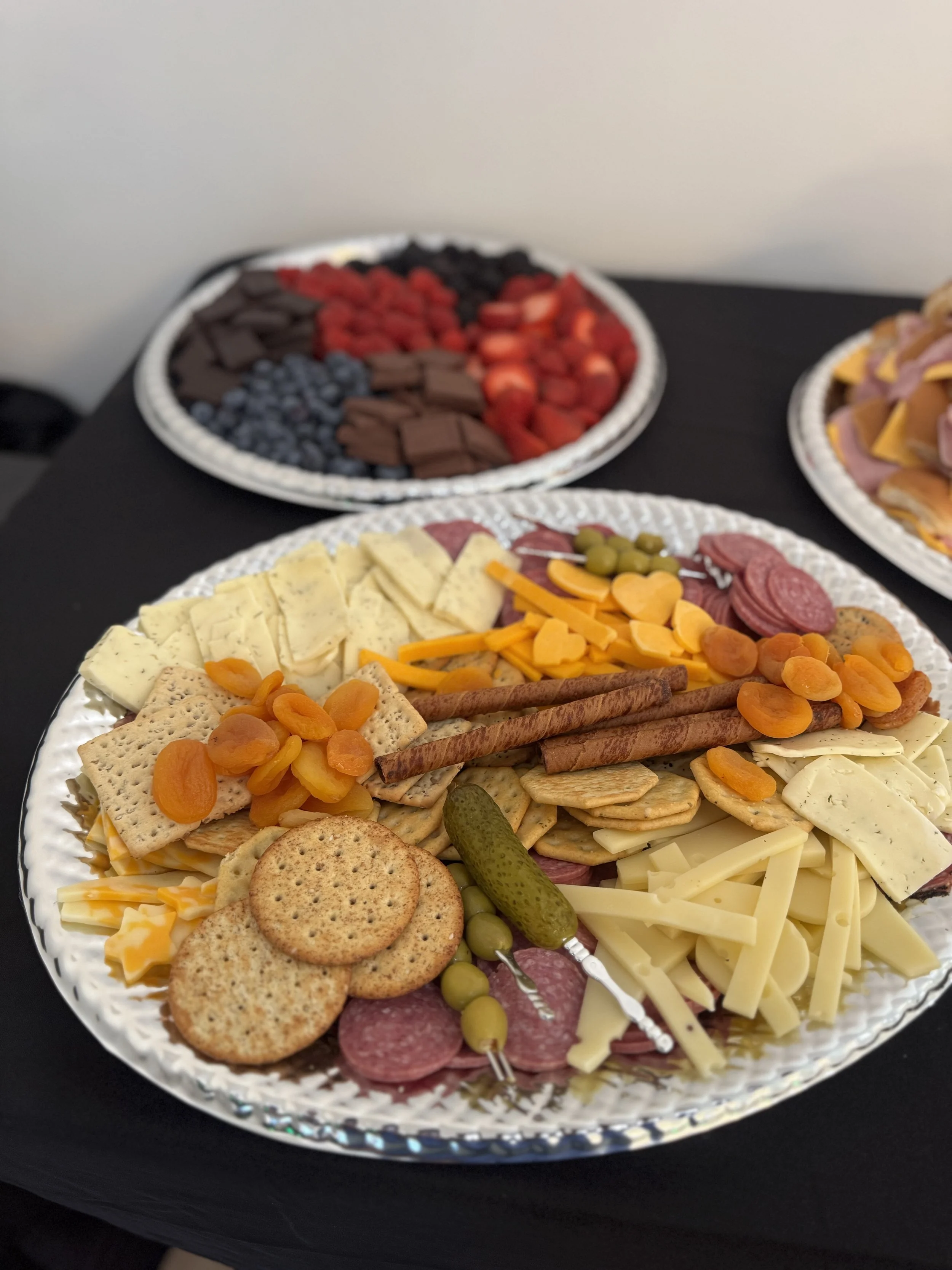 Assorted cheeses, crackers, cured meats, pickles, and dried apricots on a decorative silver platter at a buffet table.