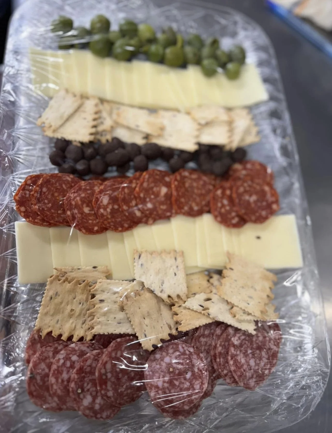 Assorted cheese, sliced salami, pepperoni, black olives, and wheat crackers on a clear plastic tray.