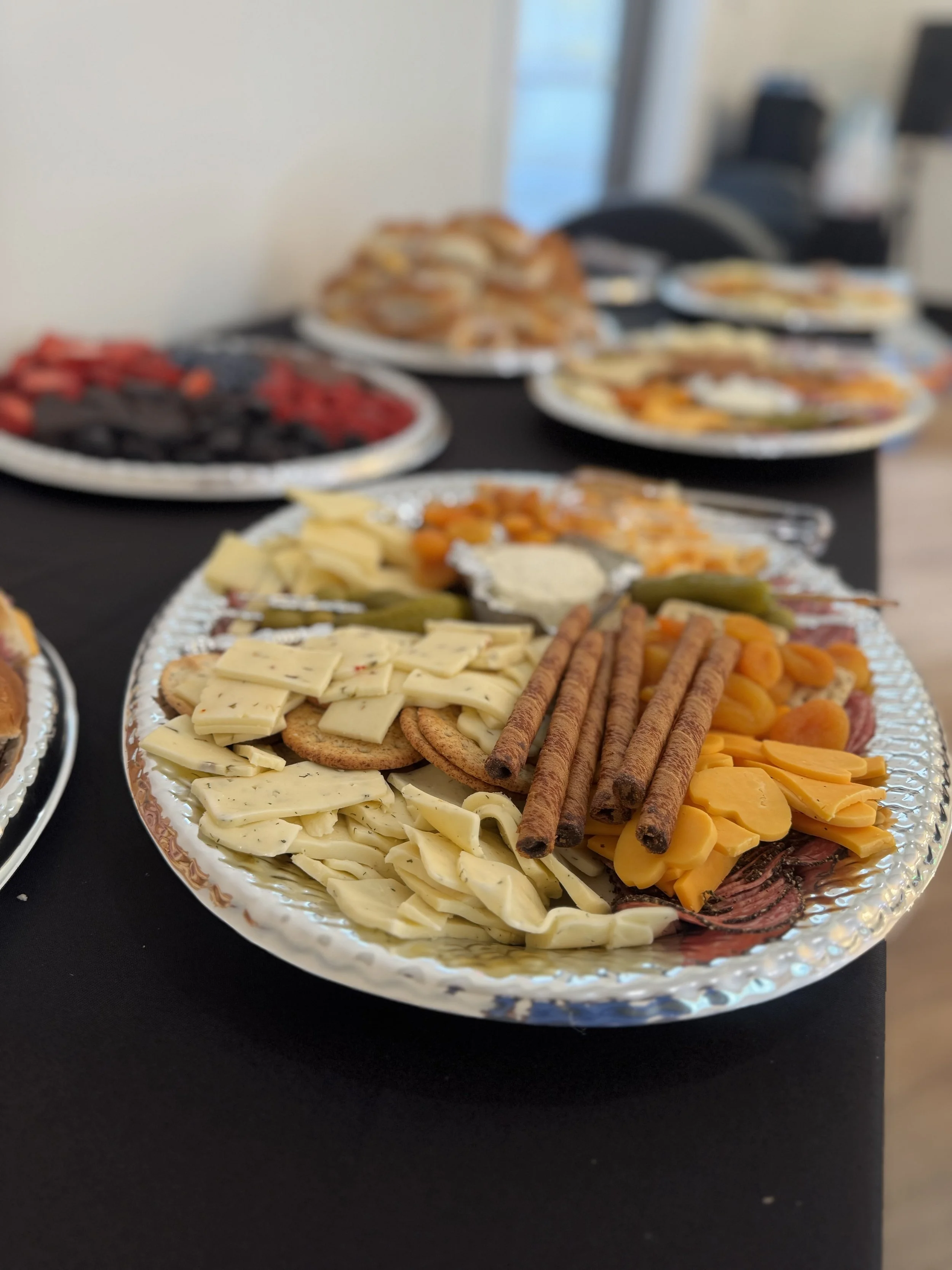 A tray of cheese, crackers, and dried sausage slices at a breakfast or brunch buffet. In the background, other plates of food including fruit and pastries.