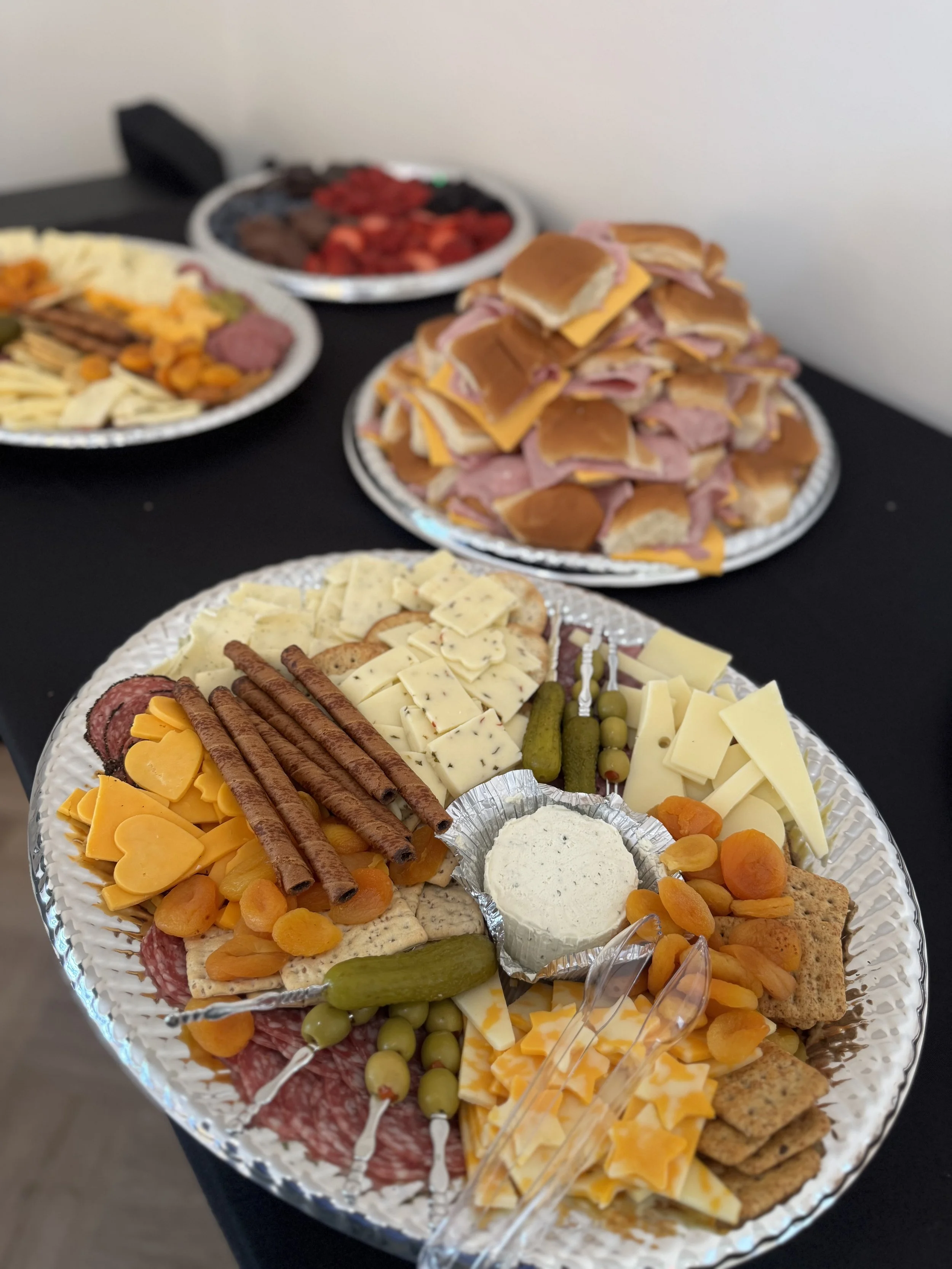 Cheese and meat platter topped with pretzel sticks, dried apricots, pickles, olives, and crackers, with other platters of sandwiches, cold cuts, and fruit in the background.