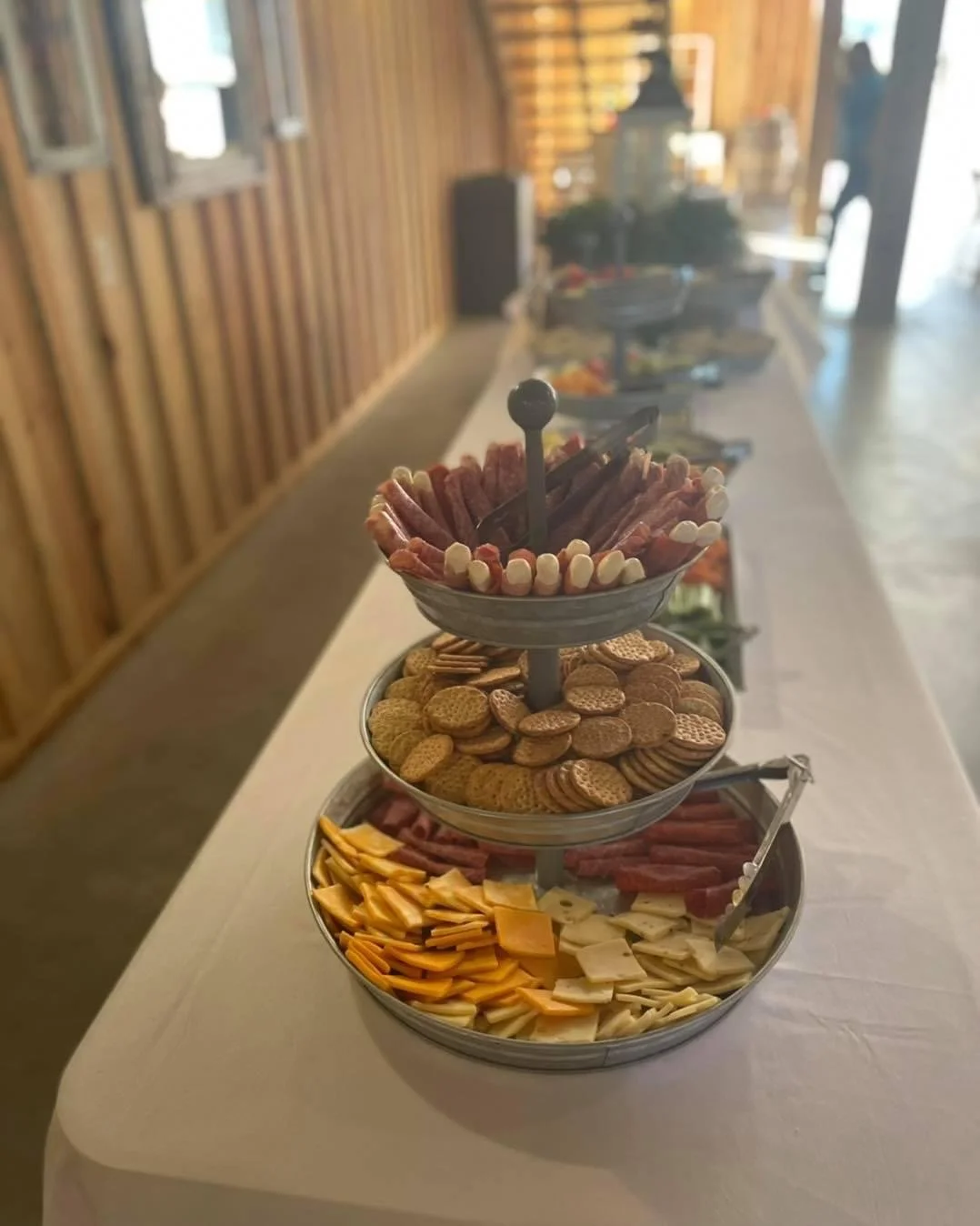 Three-tiered serving tray with assorted deli meats, cheeses, crackers, and finger foods on a white table in a rustic wooden room.
