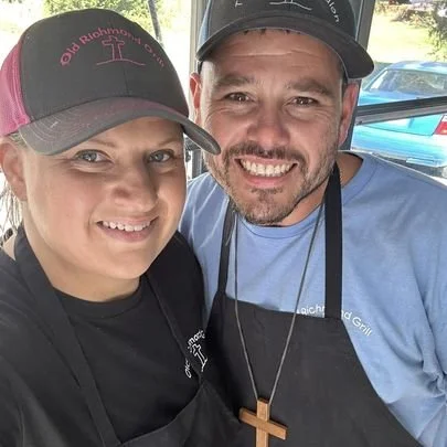 Two people smiling outdoors, wearing black hats and aprons, with a blue car in the background.