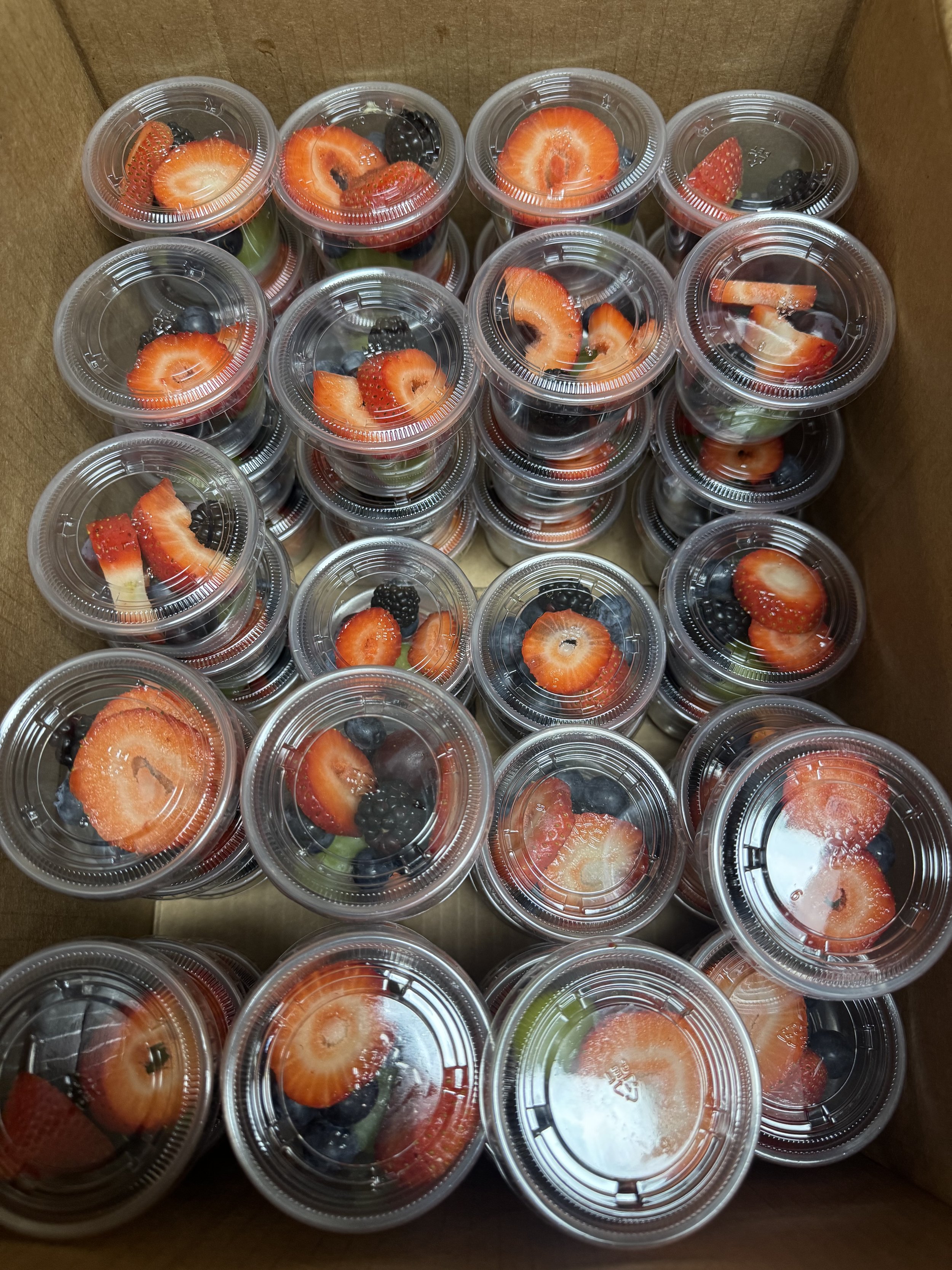 A cardboard box filled with clear plastic cups containing sliced strawberries, blackberries, and some other mixed berries.