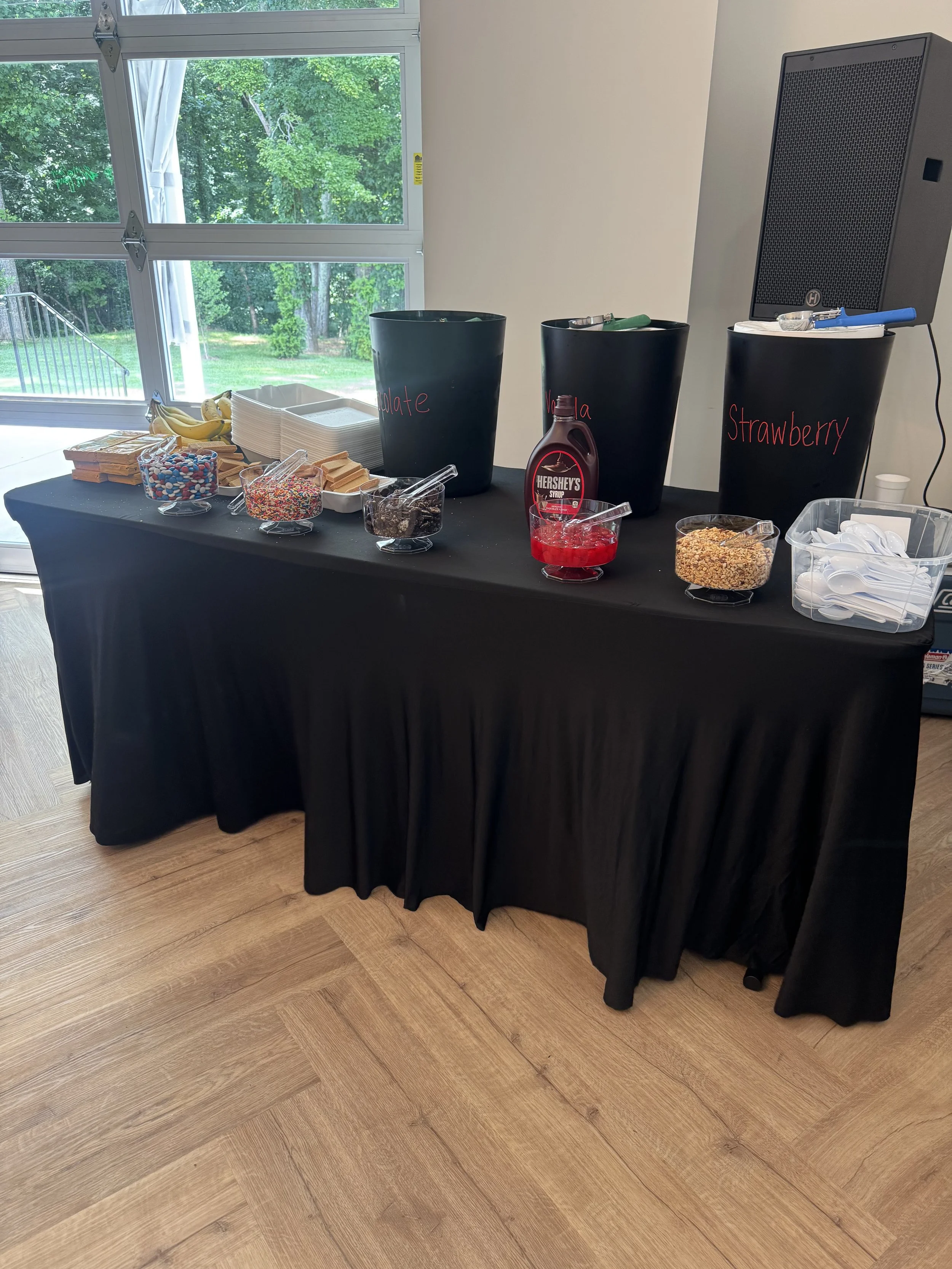 A dessert table with three large black buckets labeled 'Chocolate,' 'Vanilla,' and 'Strawberry.' The table also has bowls of sprinkles, crushed cookies, syrup, and cereal, with utensils and napkins, set against a window showing green trees outside.