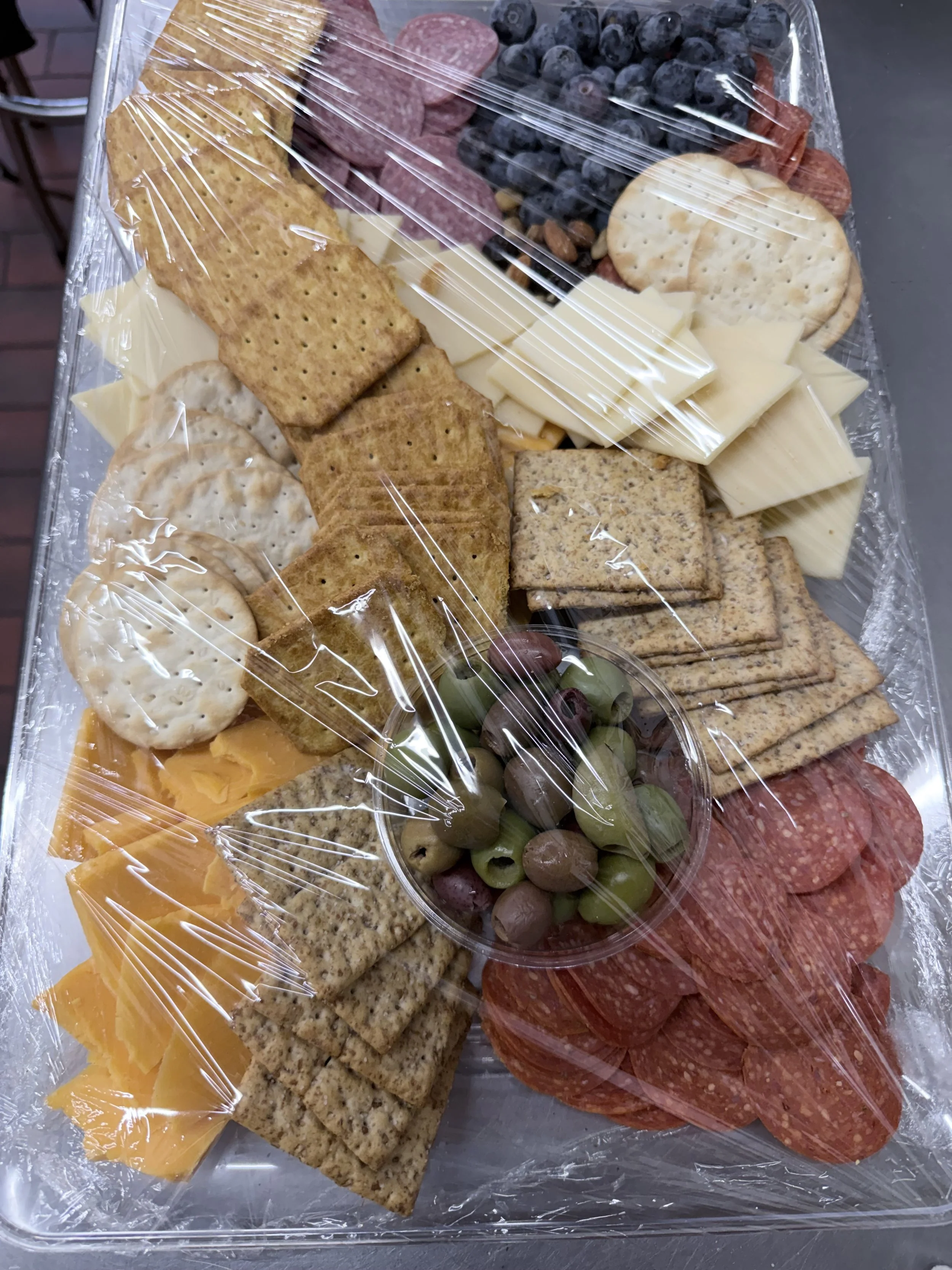 Cheese, assorted crackers, green and black olives, sliced pepperoni, sliced salami, grapes, and a mixed cheese platter covered with plastic wrap.