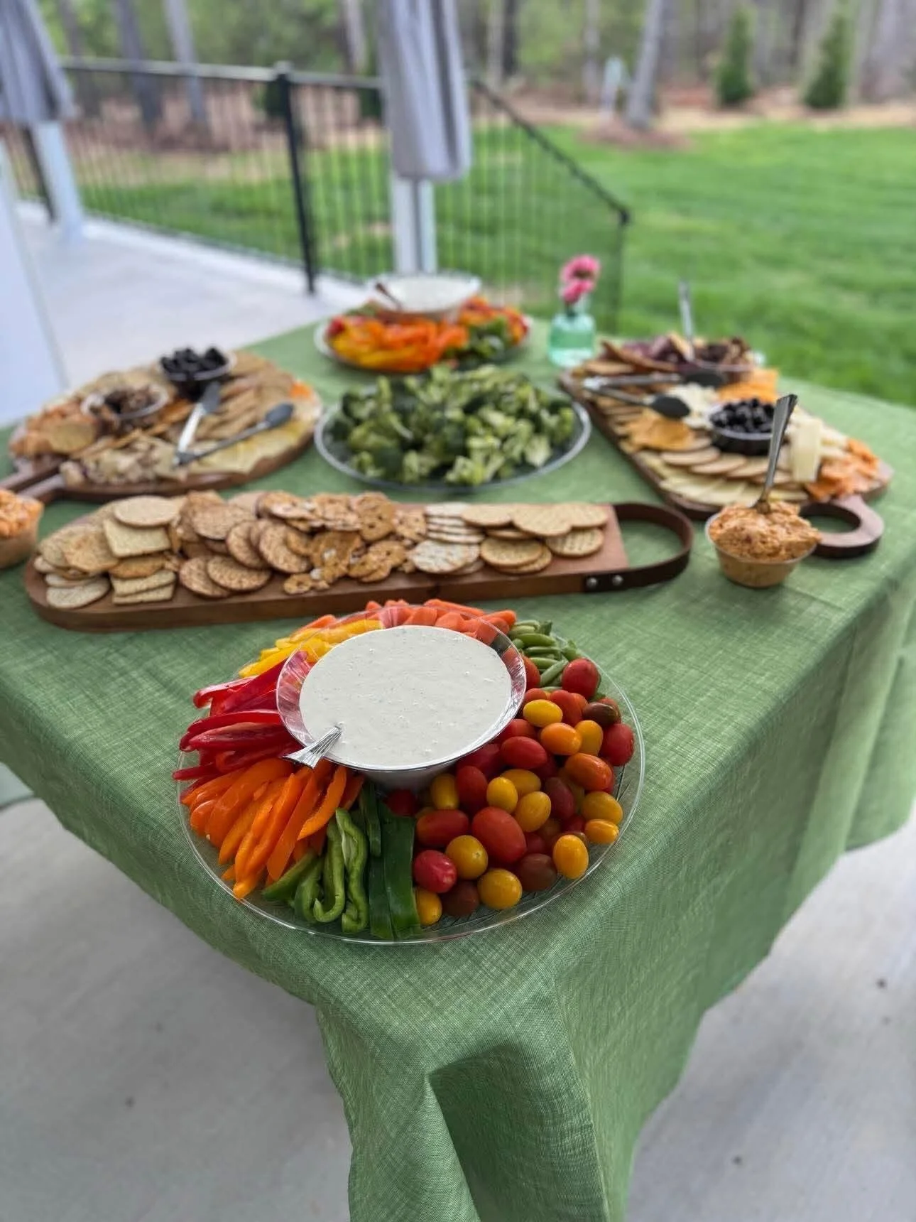 A buffet table with a green tablecloth, featuring vegetables, cherry tomatoes with dip, crackers, and an assortment of appetizers and snacks set outdoors.