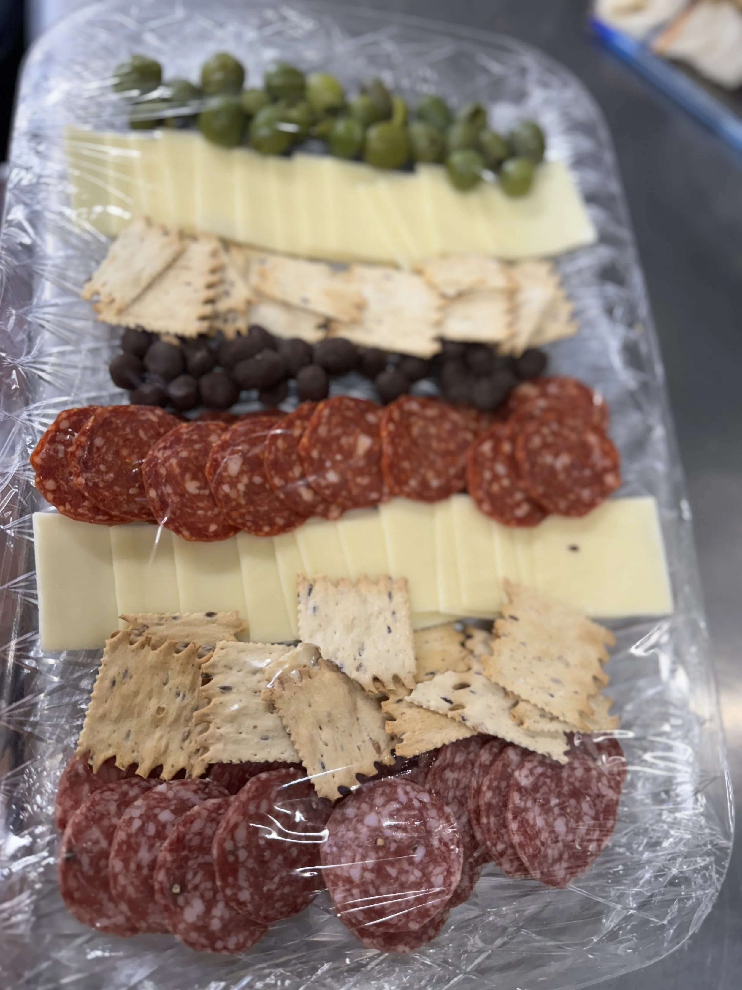 Assorted cheese and meat platter with green grapes, black and green olives, assorted sliced meats, cheese slices, crackers, and pepperoni, covered with plastic wrap.