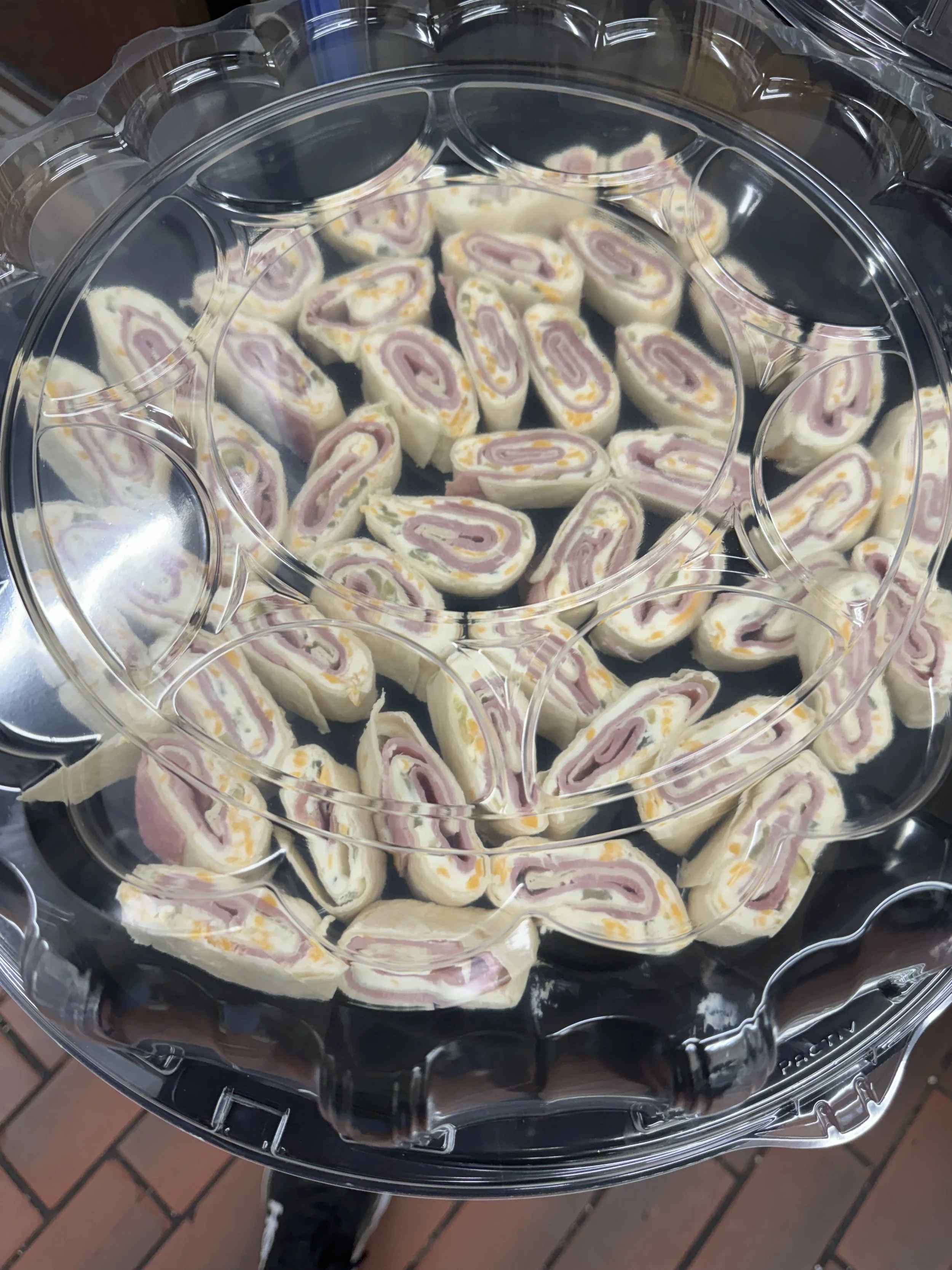 A round plastic container holding sliced pinwheel sandwiches with ham, cheese, and bread, arranged on a variety of levels.