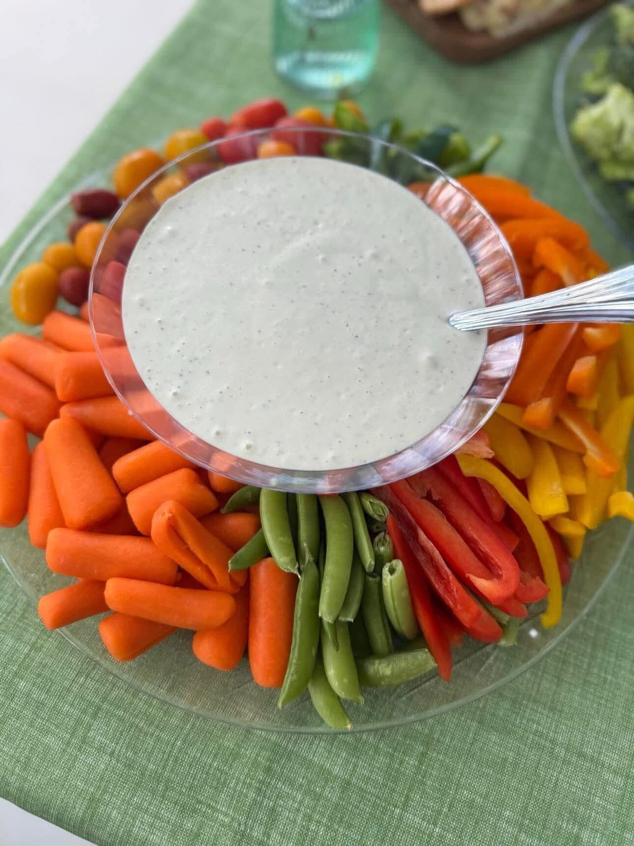 A plate of assorted raw vegetables including carrots, green beans, red and yellow bell peppers, cherry tomatoes, and grape tomatoes, with a bowl of dipping sauce on top.