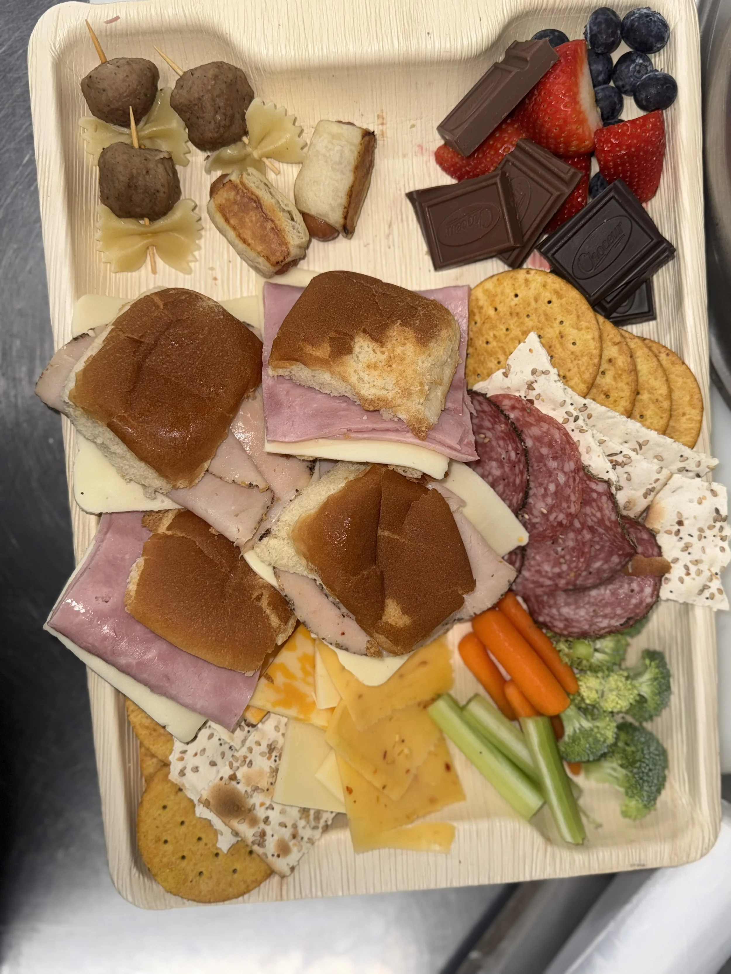 Charcuterie board with assorted meats, cheeses, crackers, strawberries, blueberries, chocolate pieces, vegetables, breaded meatballs, and small sandwiches.