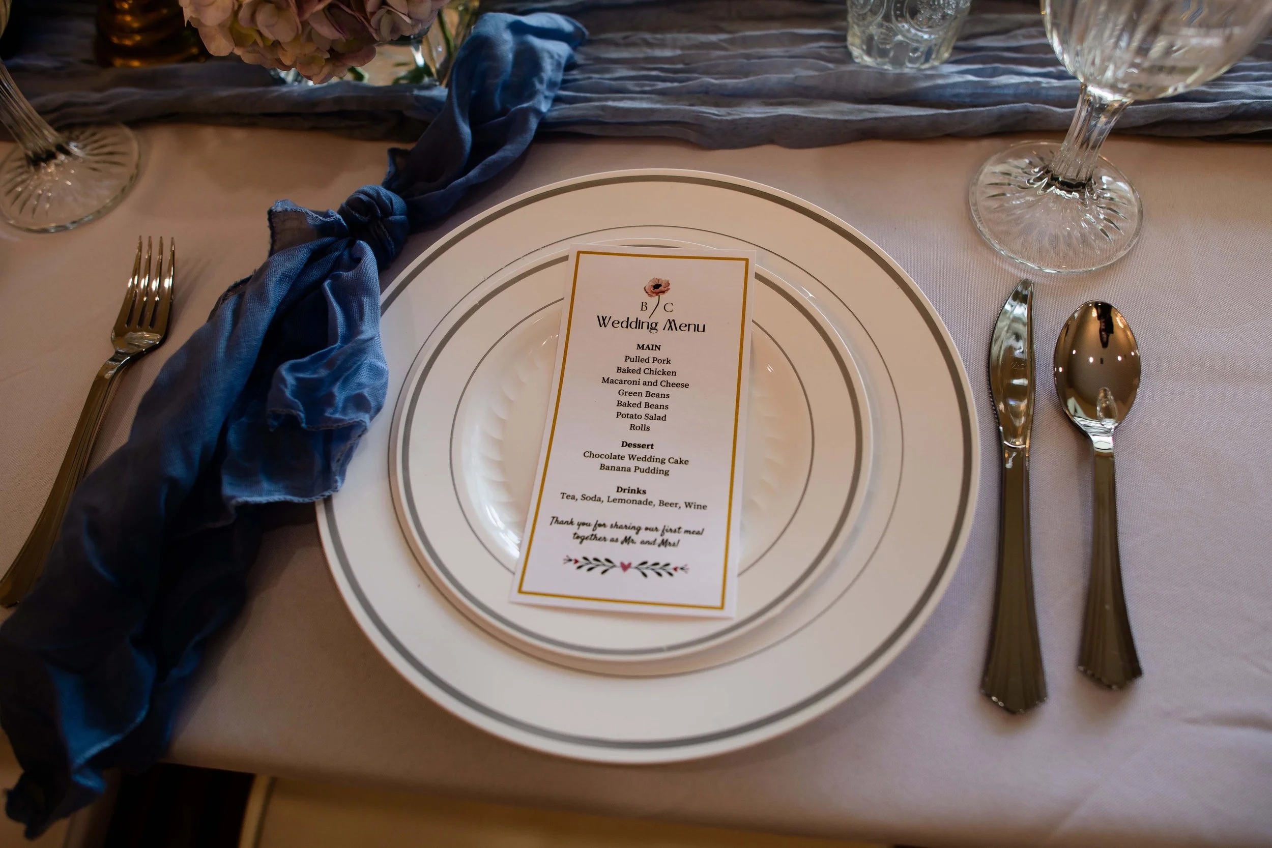 Wedding table setting with a white plate, a blue tie napkin, a wedding menu, silverware, and wine glasses on a white tablecloth.
