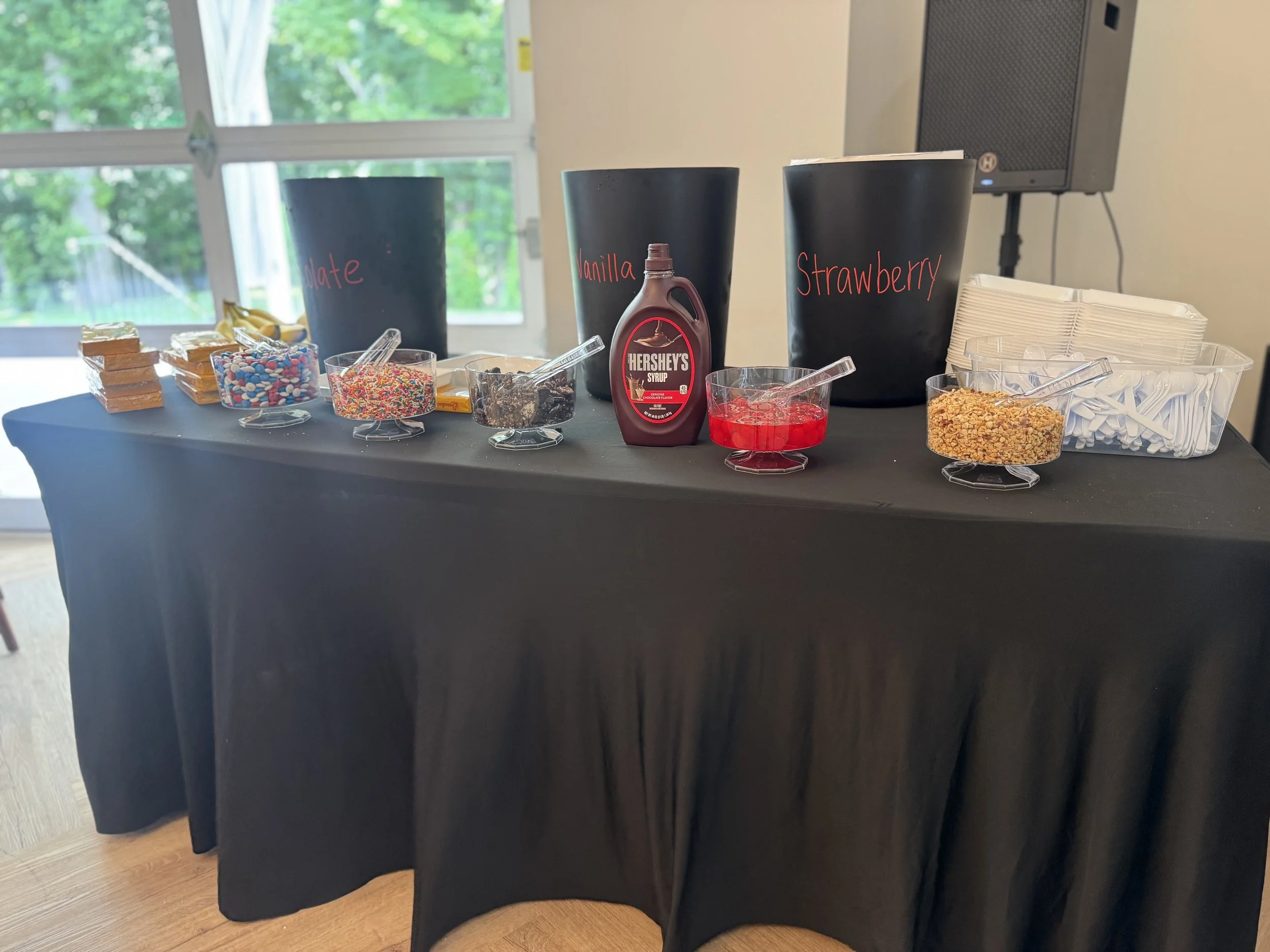 Table with various toppings in bowls labeled chocolate, sprinkles, brownie, vanilla, strawberry, crushed nuts, and a container of napkins, set up for a topping station.