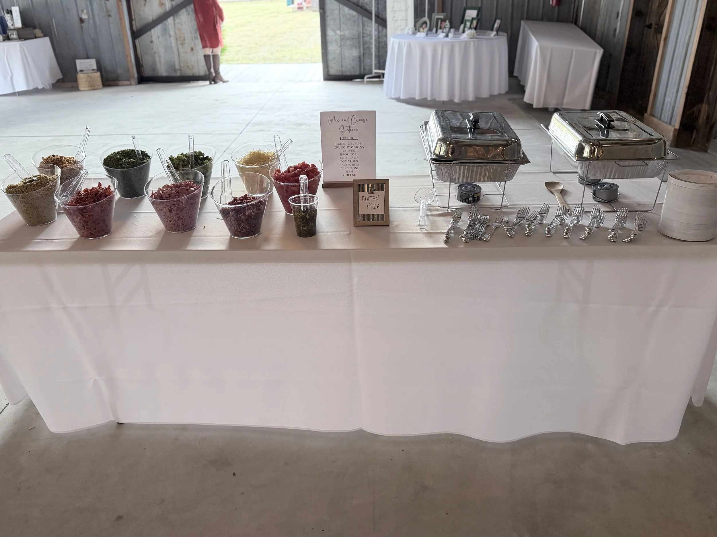 Buffet table with toppings and chafing dishes at a wedding reception, with a person standing outside the venue near open door.