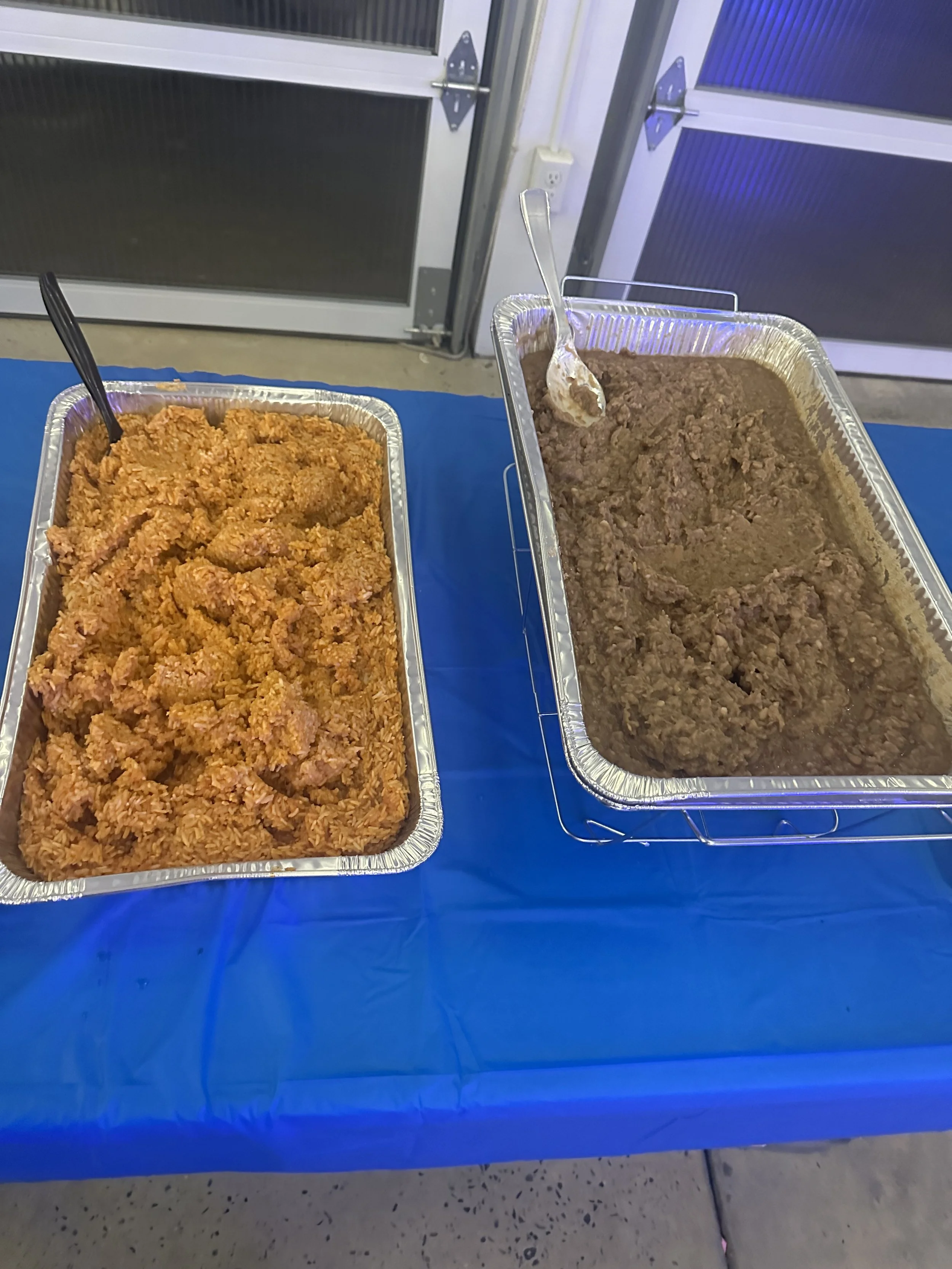 Two aluminum trays on a blue tablecloth, one with orange-colored fried rice and the other with cooked ground beef, each with a serving spoon.