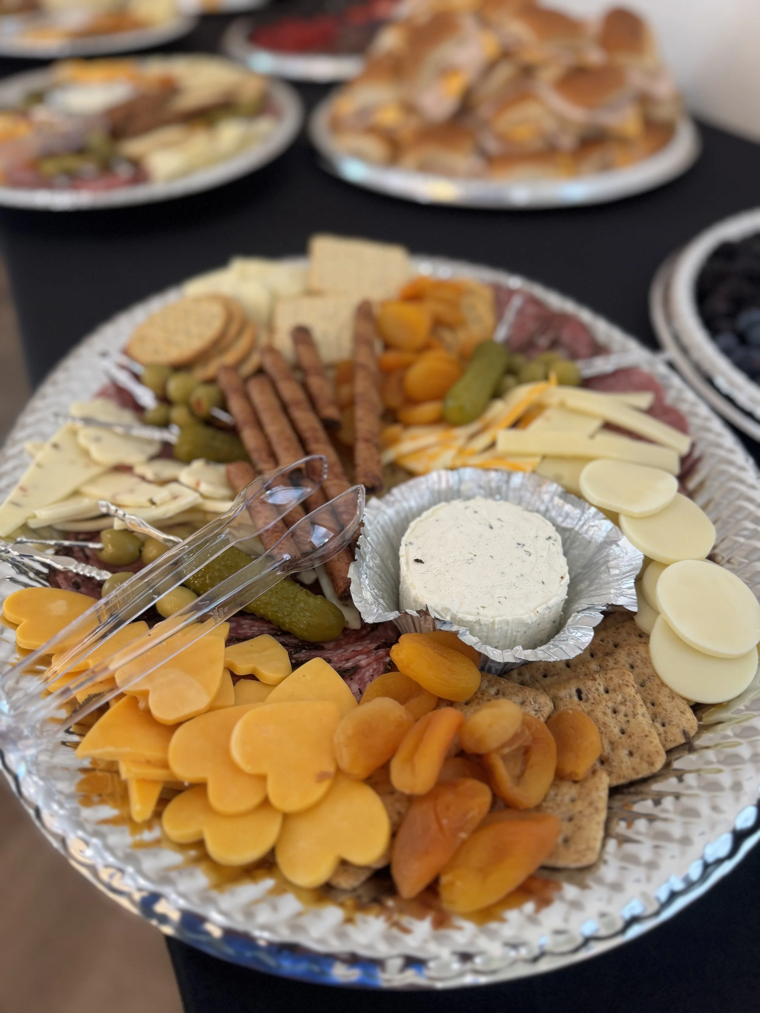 A large platter of assorted cheeses, crackers, grapes, pickles, and cured meats, with a small container of cheese spread in the center, surrounded by other platters of food on a black table.