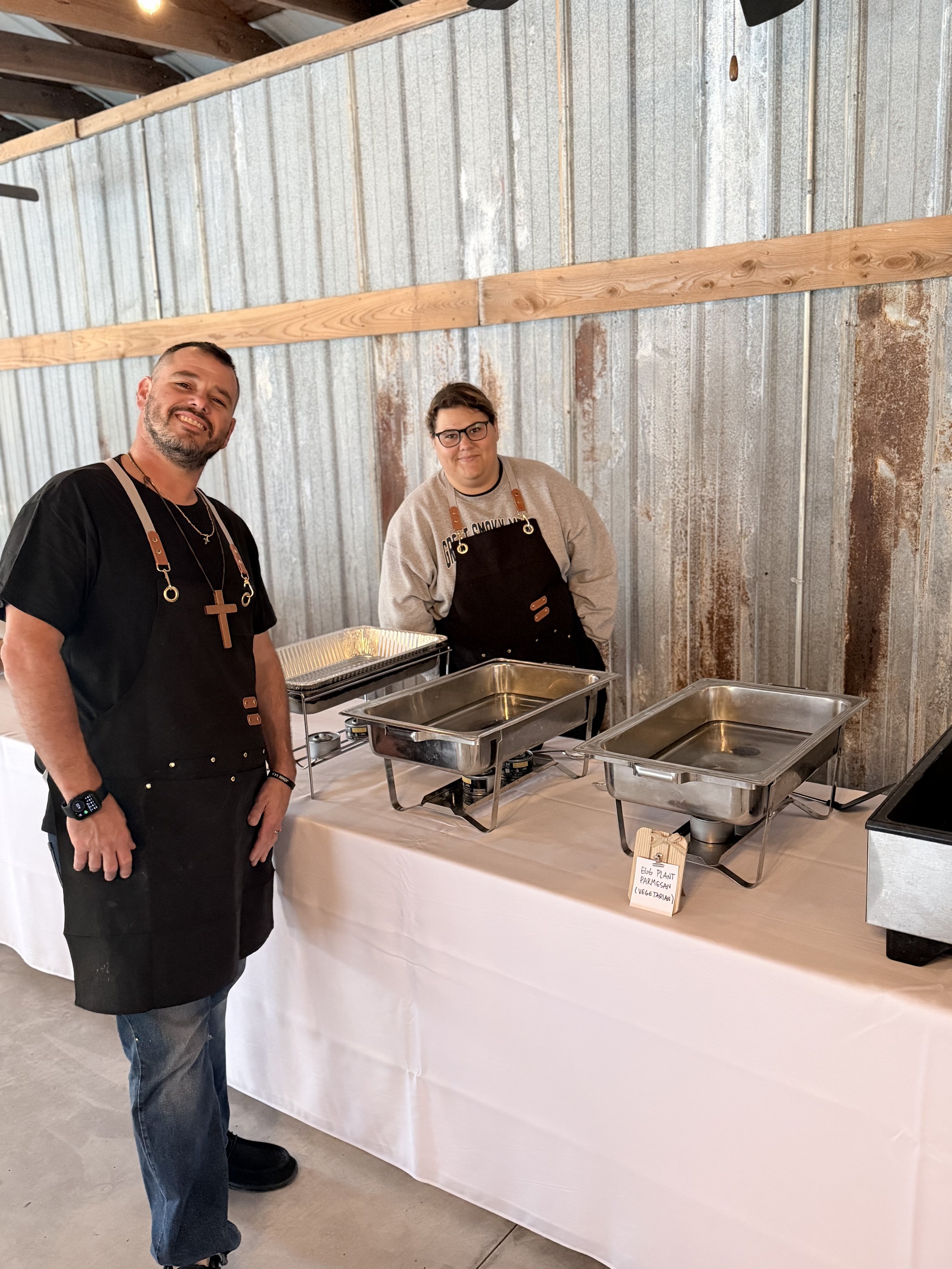 Two men standing behind a serving table with chafing dishes at an indoor event. One is smiling and wearing a black shirt and apron, the other is standing with hands in pockets and wearing glasses, a gray sweatshirt, and an apron. There is a small sig