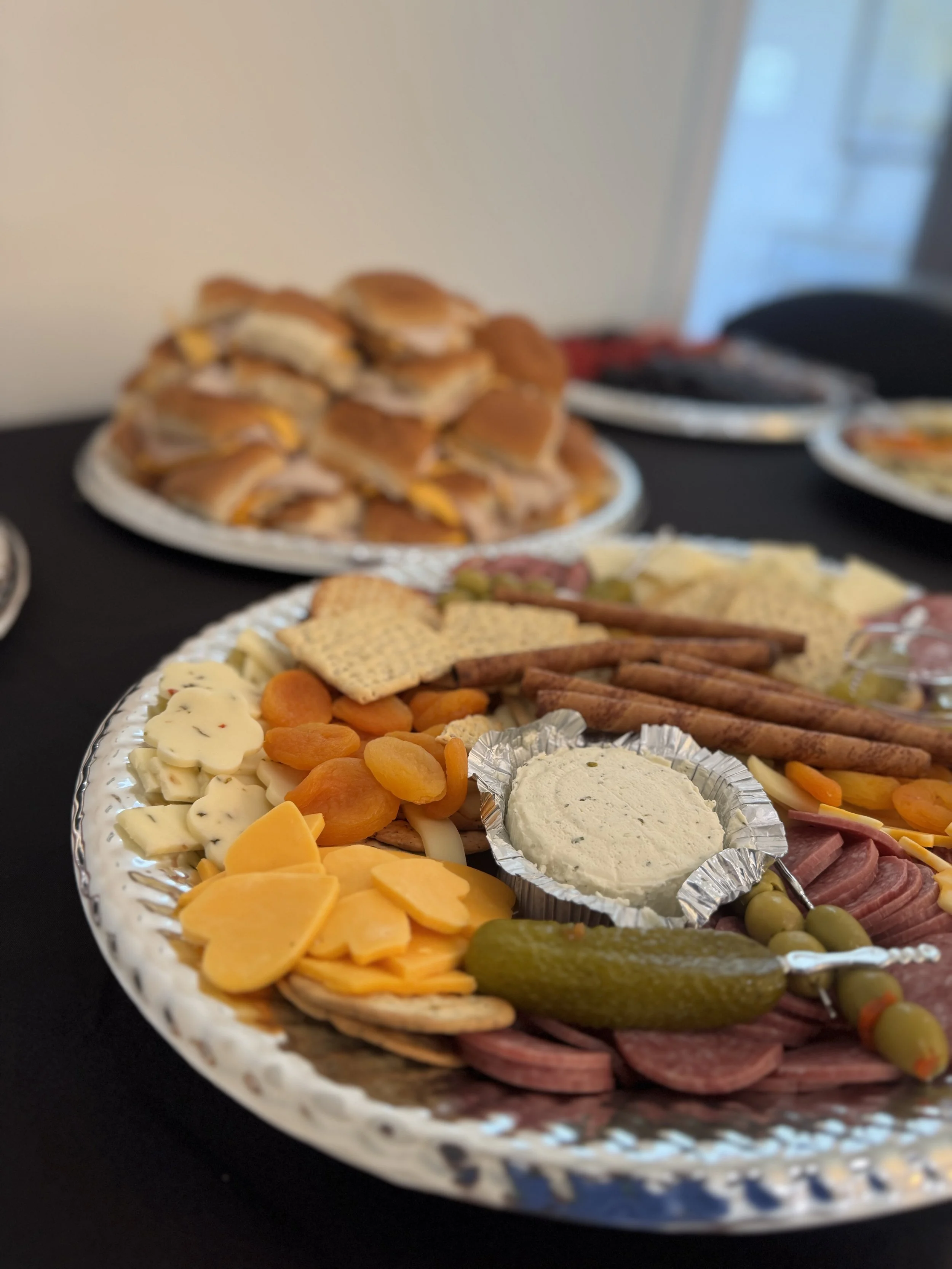 Close-up of a platter with cheeses, sliced meats, crackers, dried apricots, a pickle, and creamy dip, with sandwiches in the background.