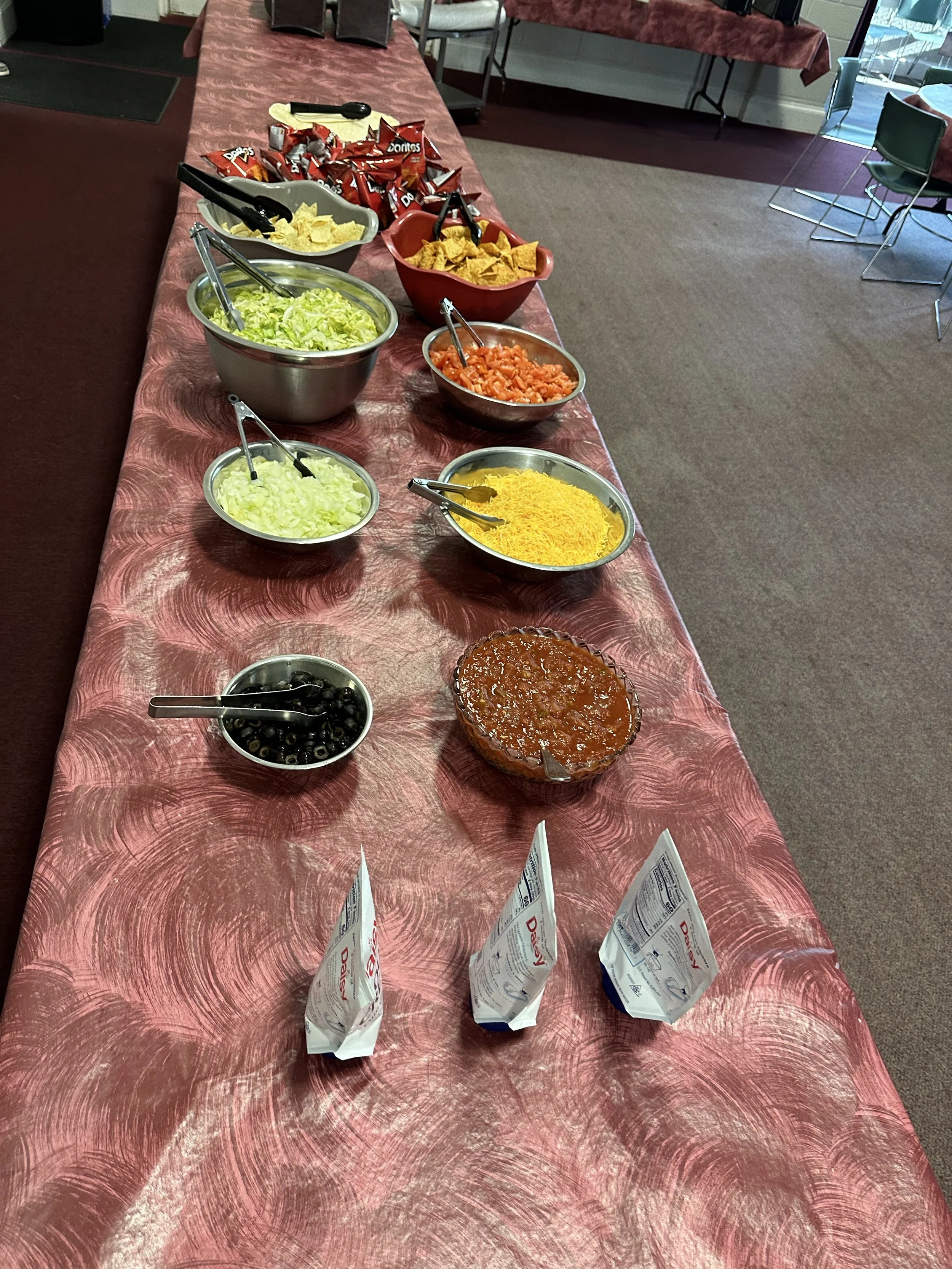 A buffet table with various toppings including shredded cheddar cheese, black olives, chopped tomatoes, shredded lettuce, chopped onions, shredded cheese, salsa, and bags of Doritos chips underneath the bowls, set up for a taco bar.