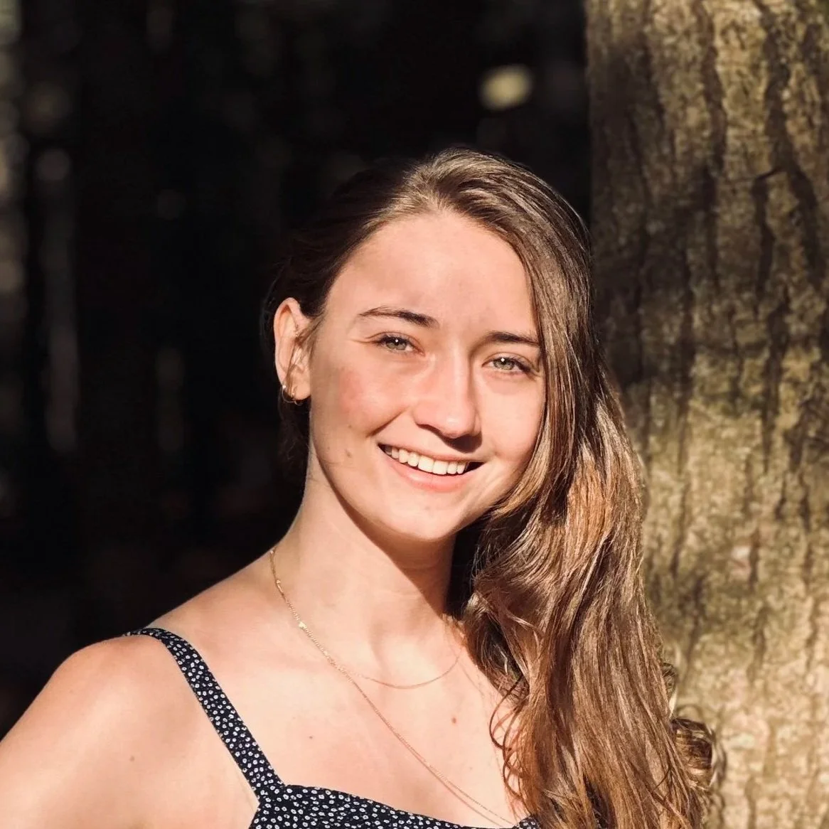 Portrait of Noemi Roestel smiling outdoors, standing next to a tree in natural sunlight.