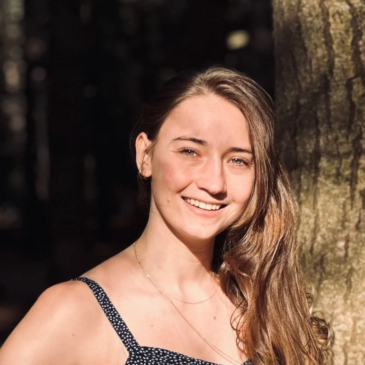 Portrait of Noemi Roestel smiling outdoors, standing next to a tree in natural sunlight.