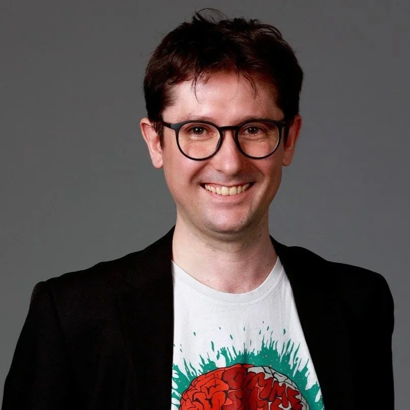 Portrait of Guillaume Dumas smiling, wearing glasses, a black jacket, and a printed T-shirt, with a neutral background.