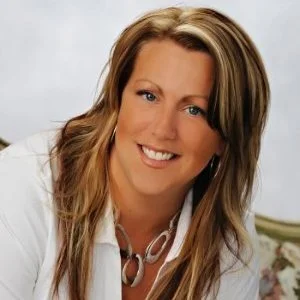 Portrait of Dominique Chabot with long blonde hair, smiling and wearing a white top, photographed indoors against a light background.