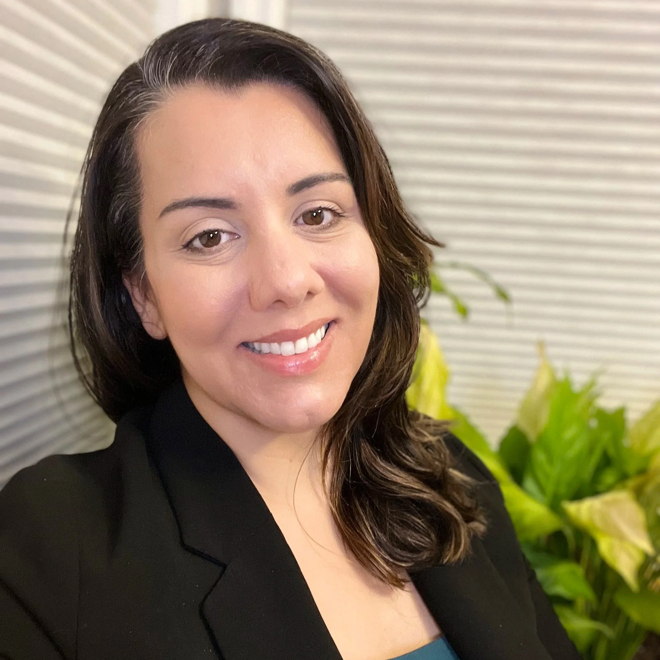 Portrait of Sarah Banner smiling, wearing a teal blouse and dark blazer, with green plants and blinds in the background.