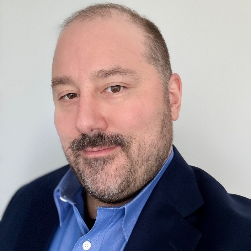 Portrait of Daniel Nerenberg smiling slightly, wearing a blue shirt and dark blazer, with a light neutral background.