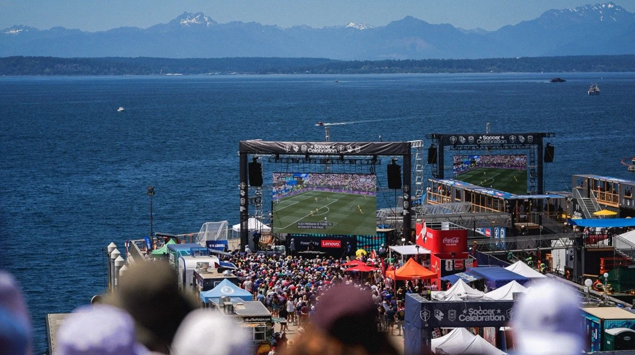 Photo of a crowd watching a soccer match on Pier 62 with the barge, water and mountains in the background