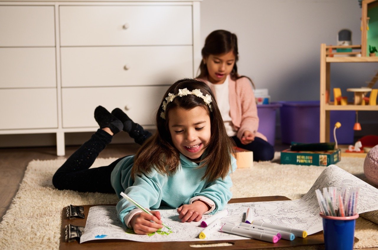 Young girl laying on the floor coloring with another child in the background