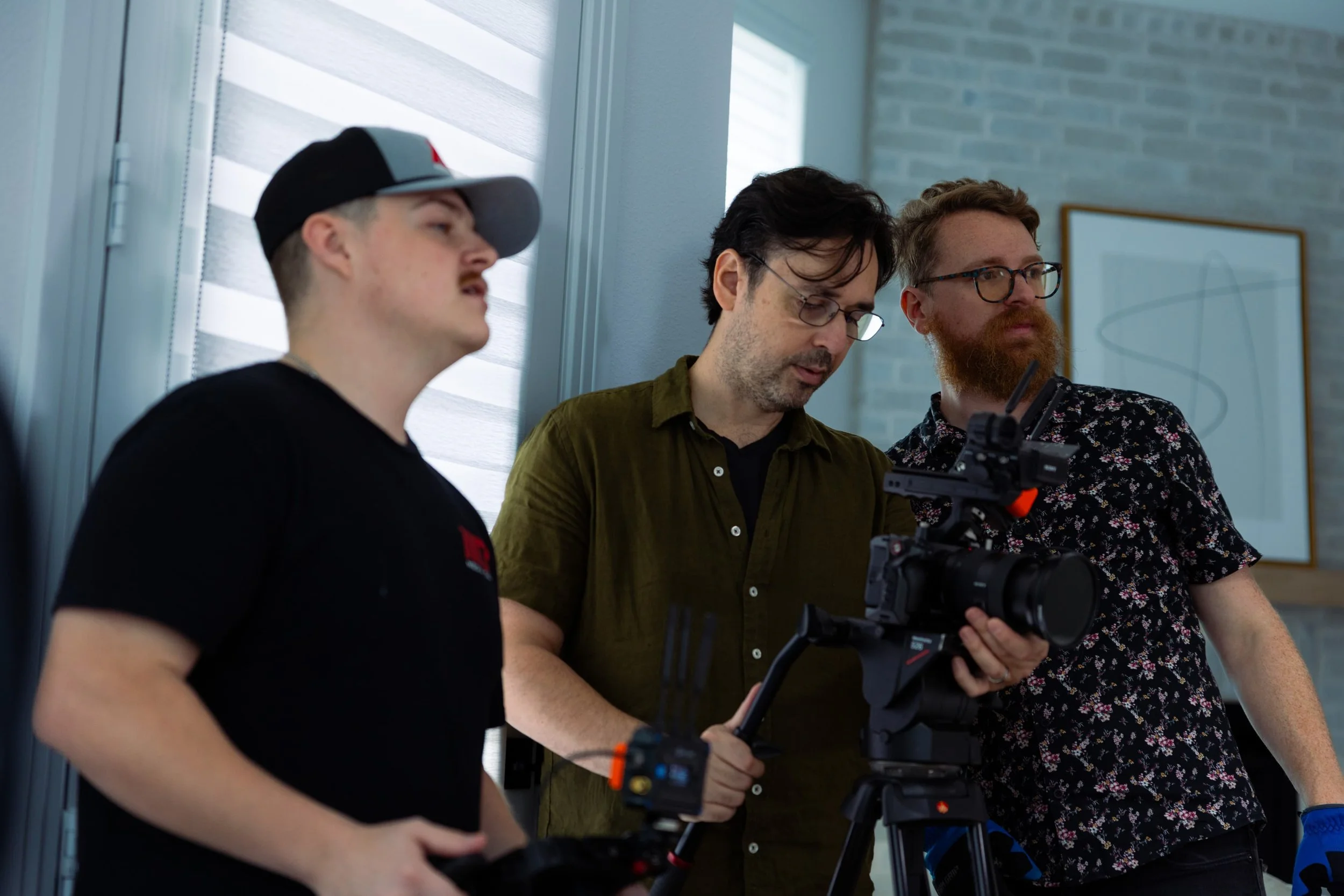 Three men working with film or video equipment indoors, near a window with blinds, with a brick wall and framed art in the background.