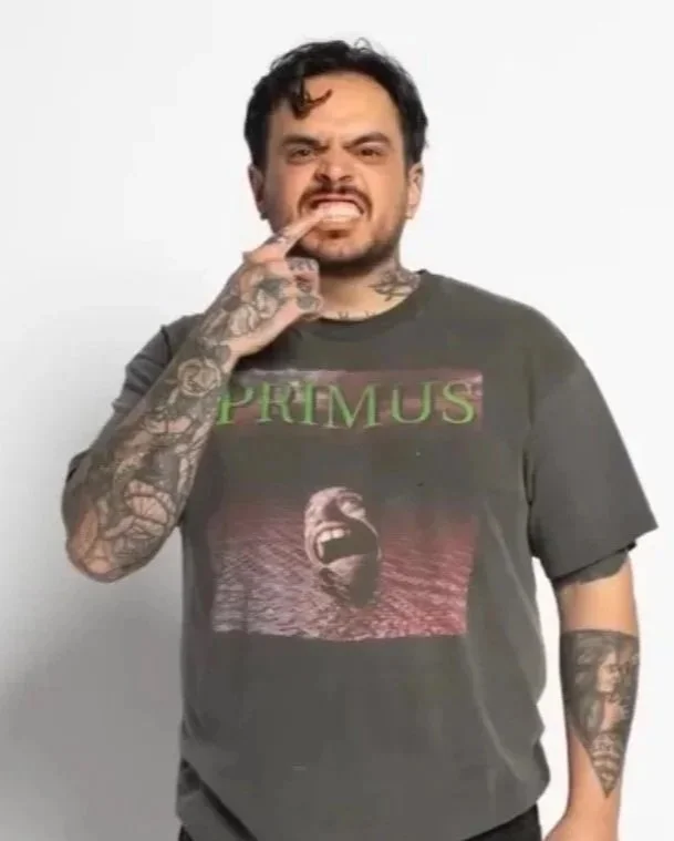 A man with dark hair and tattoos, wearing a Primus band T-shirt, makes a grimacing expression while touching his lips with his finger.