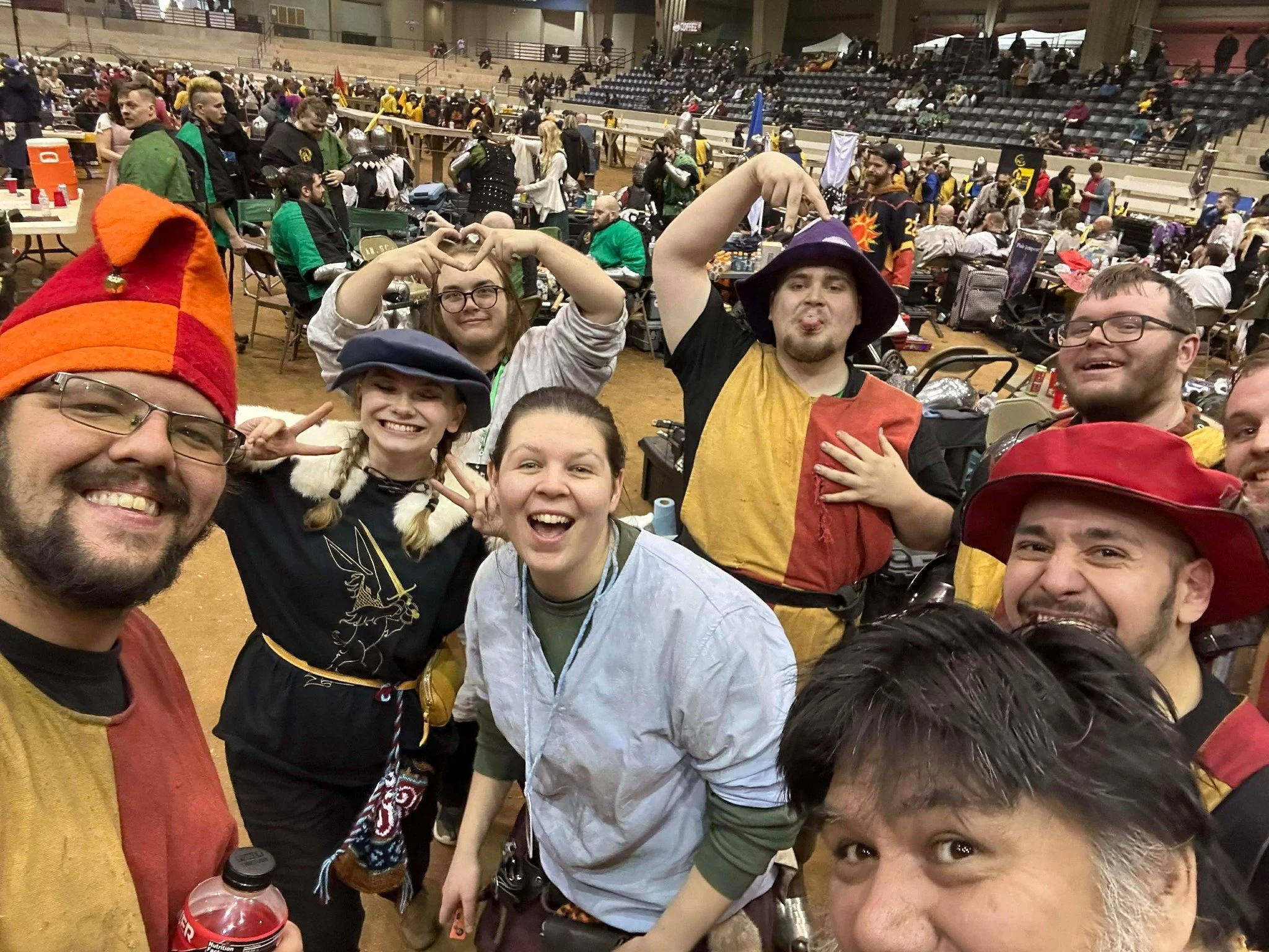 A group of smiling people dressed in medieval and fantasy costumes posing for a selfie at a large indoor event with crowded seating and costumed attendees in the background.