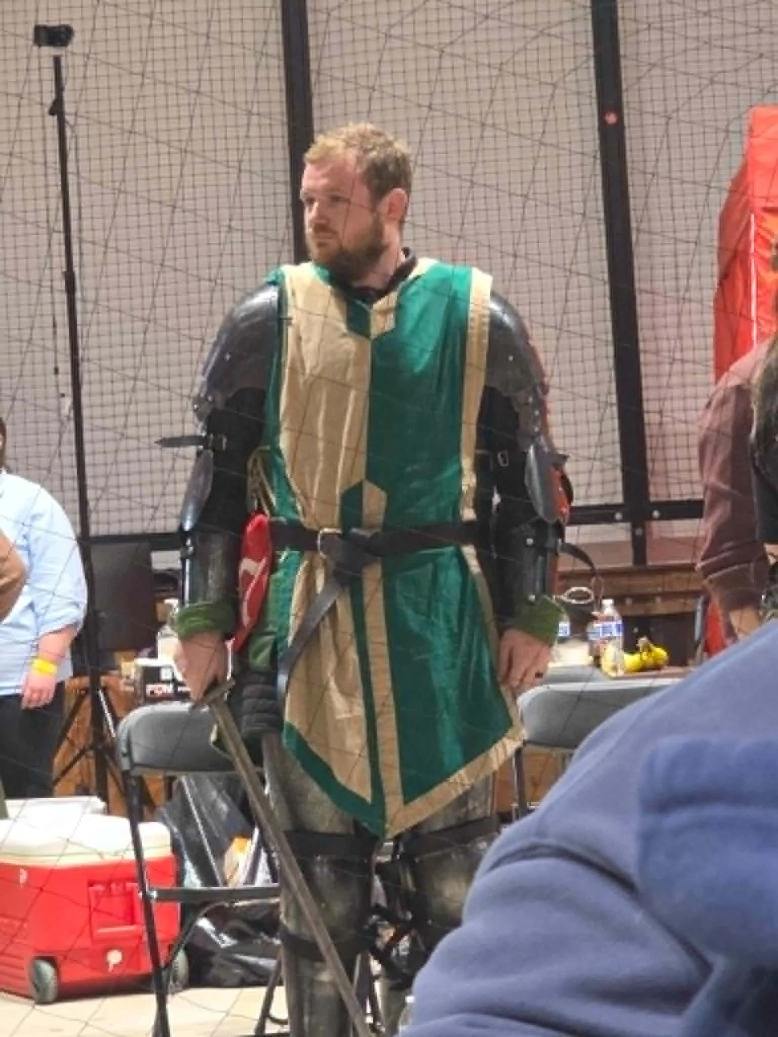 A man dressed as a medieval knight, wearing armor and a green and gold robe, standing indoors with chairs and tables around him, and a netted barrier in the background.