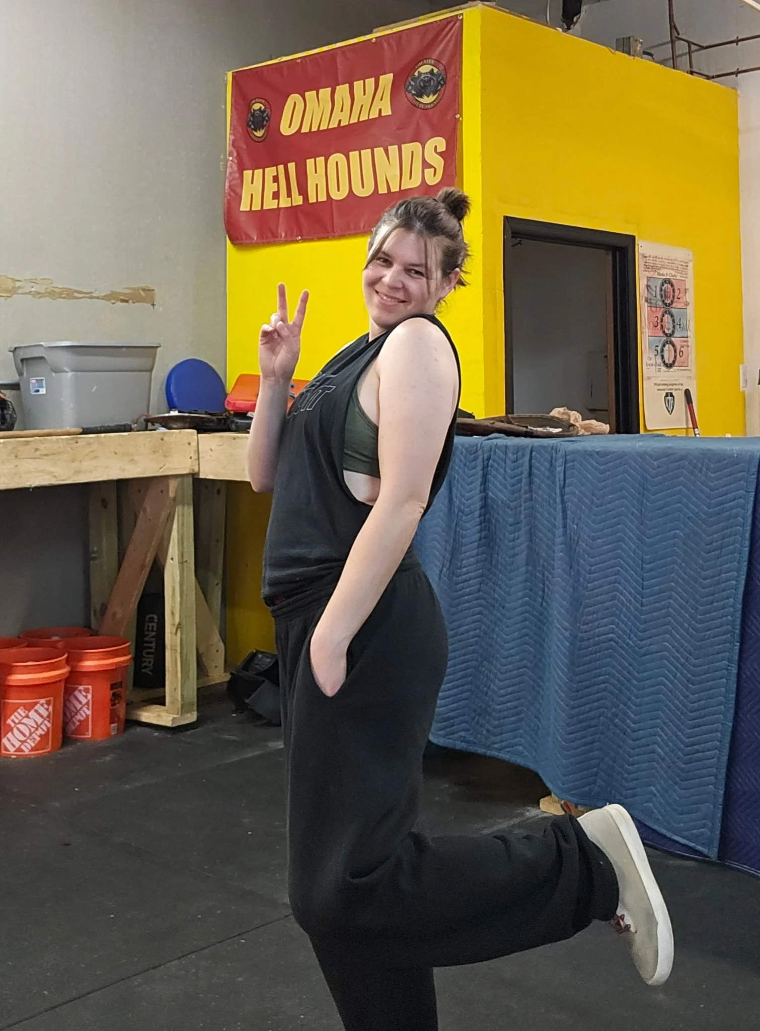 A woman with brown hair tied in a bun, smiling and making a peace sign with her hand, while kneeling on one knee in a gym. She is wearing a black sleeveless top, black pants, and white sneakers. She is standing in front of a yellow wall with a red banner that reads 'OMAHA HELL HOUNDS' and logos of a black hound's head. To her left are orange buckets labeled with The Home Depot logo and a wooden workbench. To her right, there is a blue padded mat and a doorway.