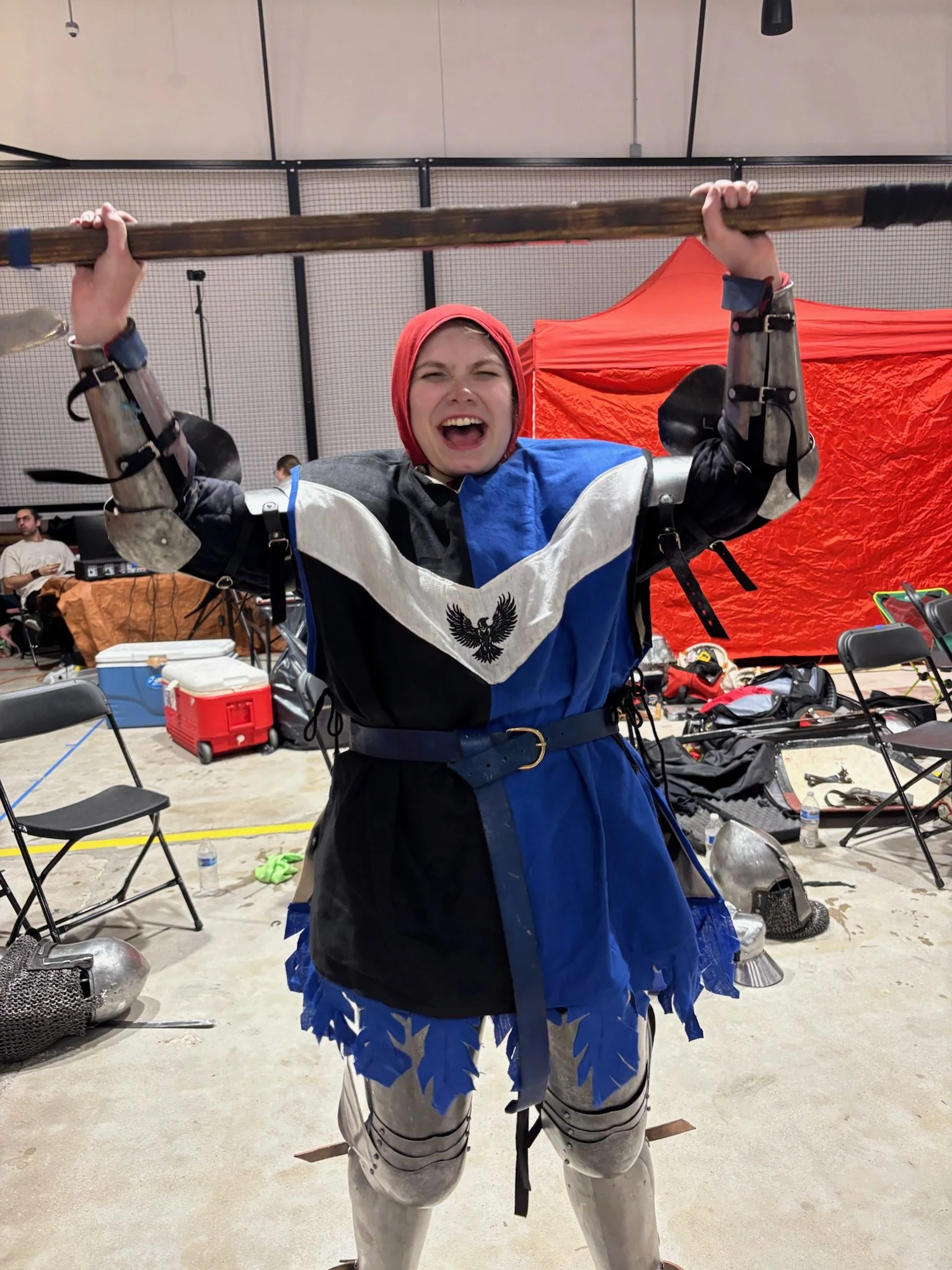 Person wearing medieval armor and a red hood, holding a wooden sword above their head, in an indoor setting with chairs, tables, and equipment visible in the background.