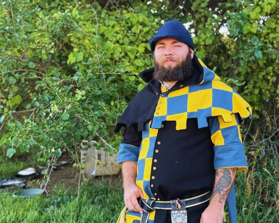 A man with a beard and tattooed right arm stands outdoors in front of green foliage, wearing a medieval-inspired costume with a blue and yellow checkered pattern, a black shirt, and a matching hat.