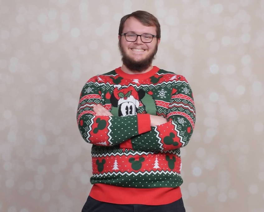A smiling man with glasses, a beard, and a holiday sweater with festive patterns and Disney characters, standing with crossed arms in front of a sparkly holiday-themed background.