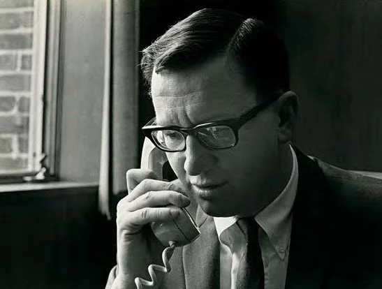 Man with glasses talking on a landline phone in a black and white photo.